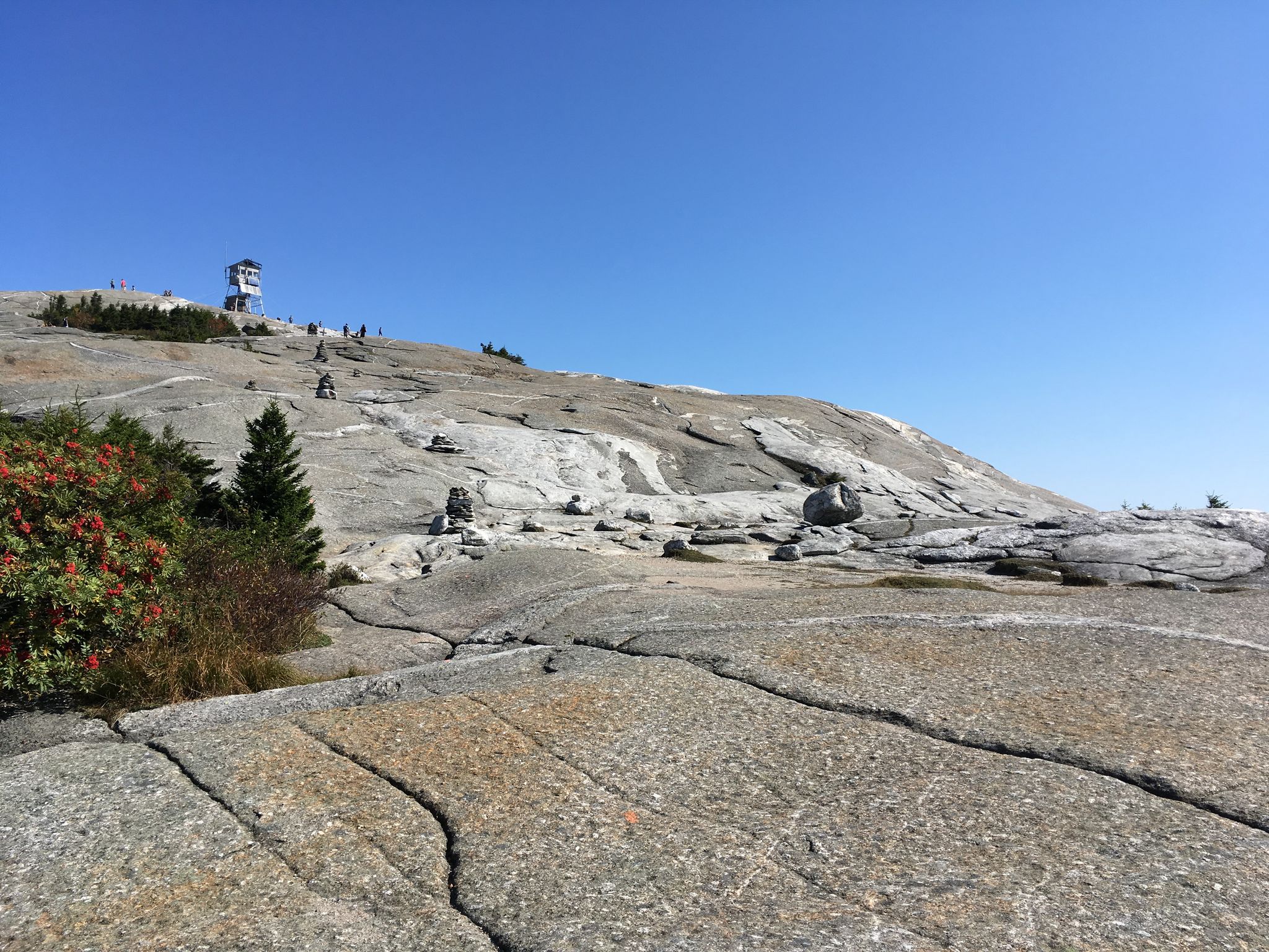 Rocky mountain summit with a distant fire tower and small figures of people. - West Ridge Trail