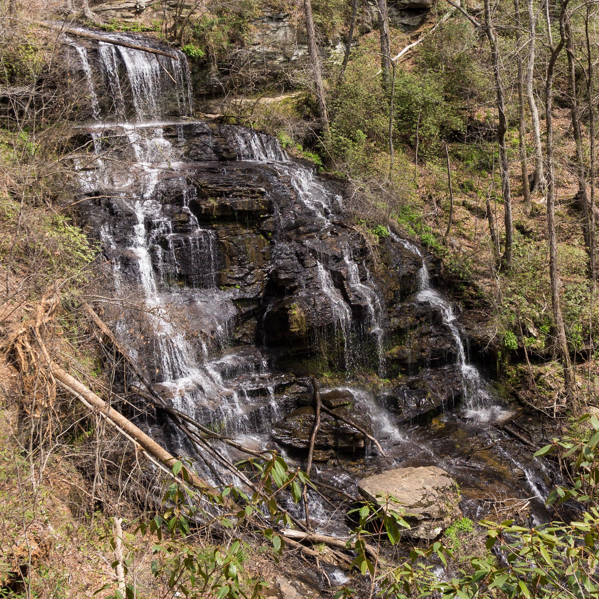 Multi-tiered waterfall cascading over dark rocks in a sunlit, wooded landscape.
