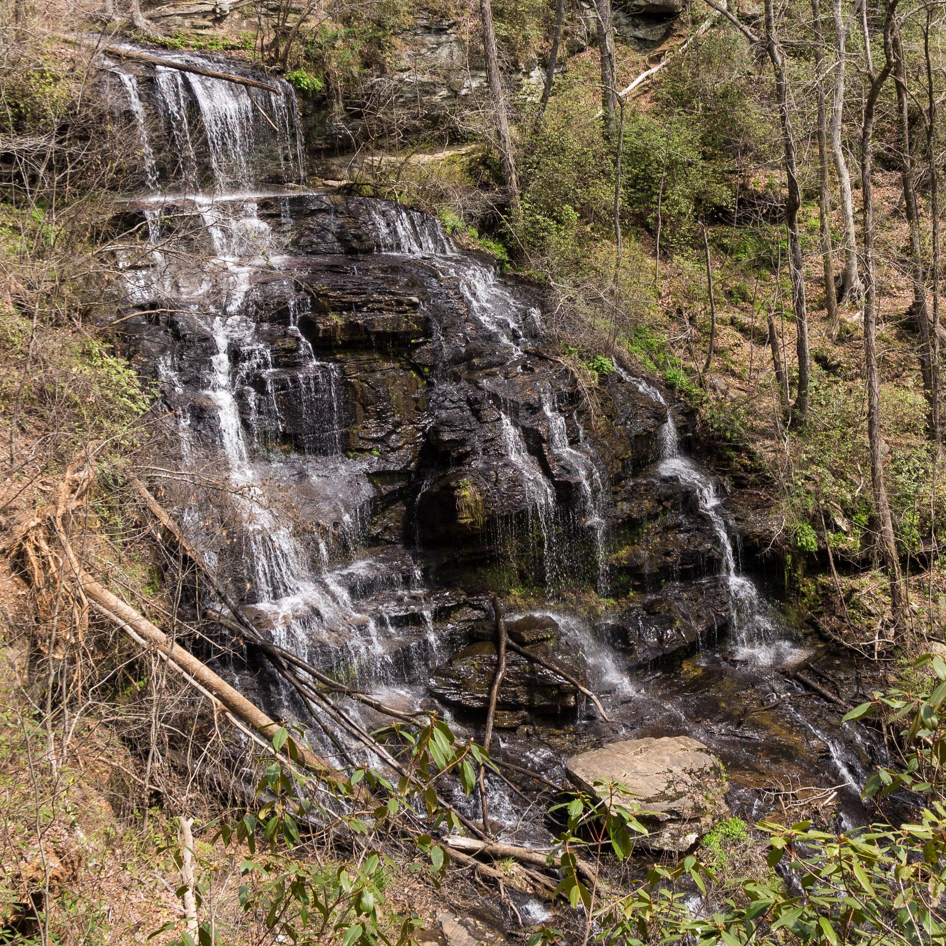 Multi-tiered waterfall cascading over dark rocks in a sunlit, wooded landscape.