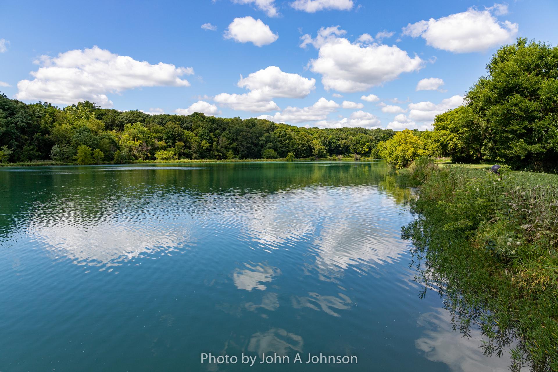 Vibrant blue lake reflecting clouds, surrounded by lush green trees on a sunny day.