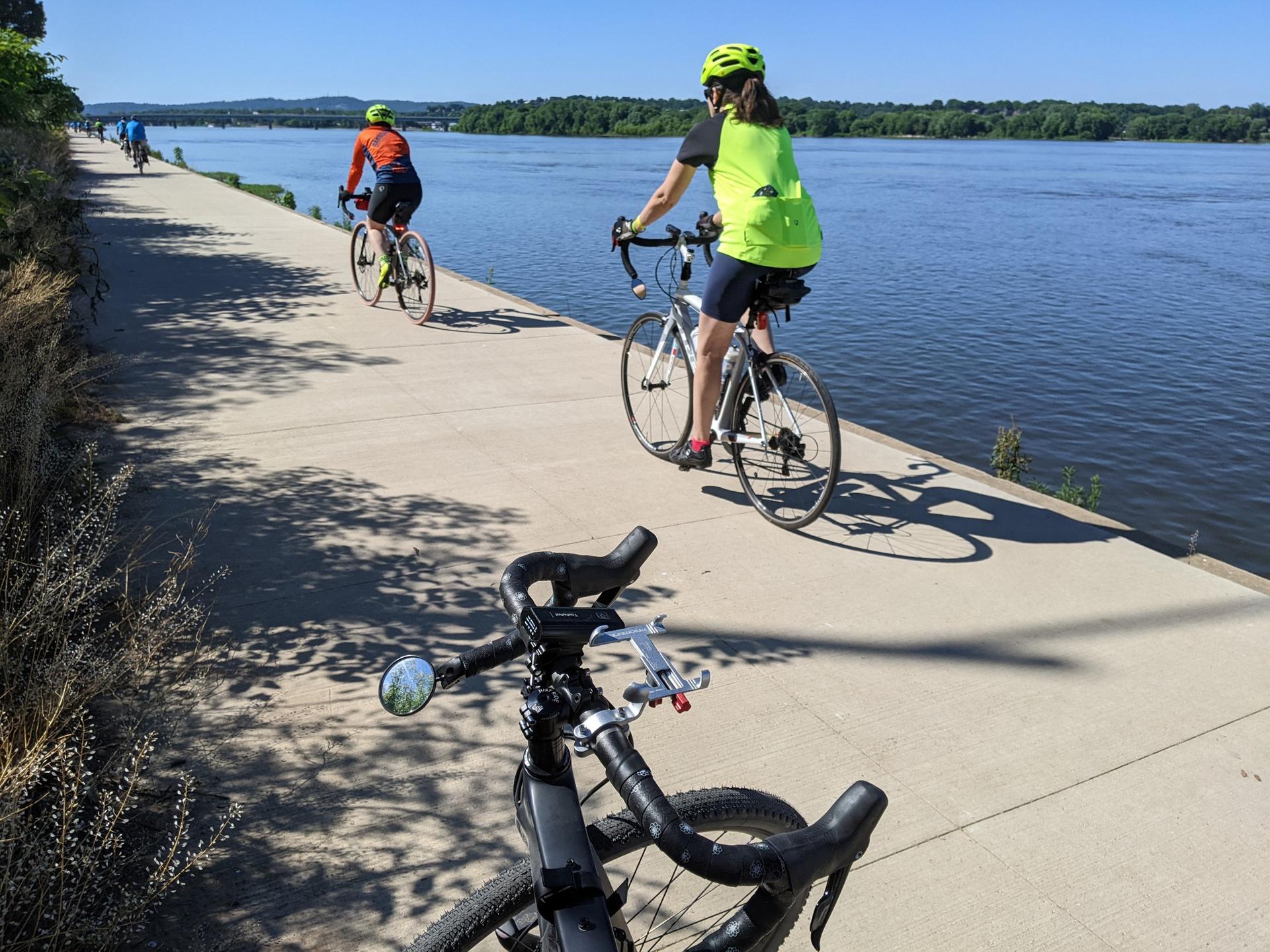 Two cyclists ride on a paved path next to a wide river on a sunny day.