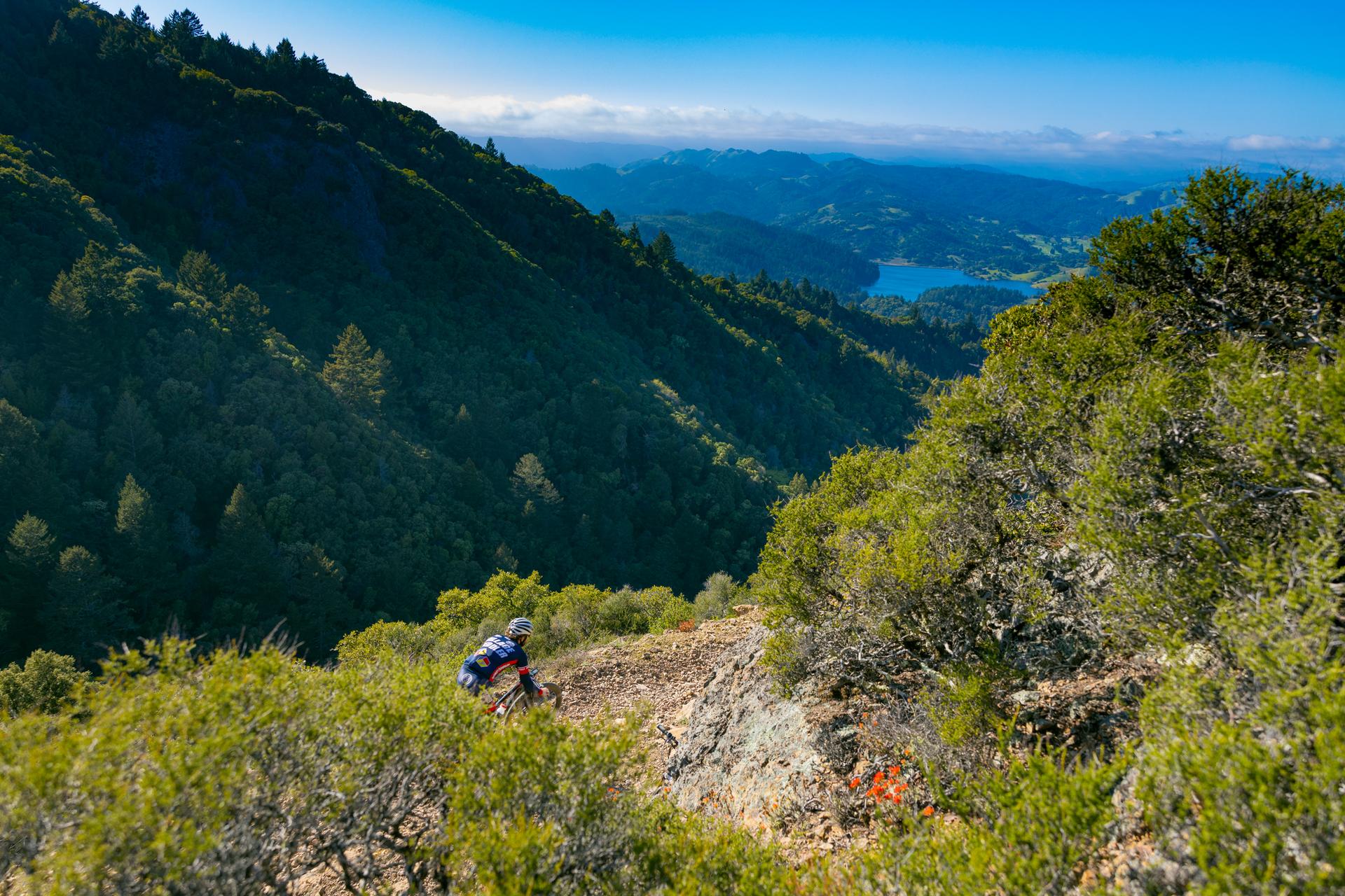 A mountain biker descends a steep, sunny dirt trail overlooking a distant lake.