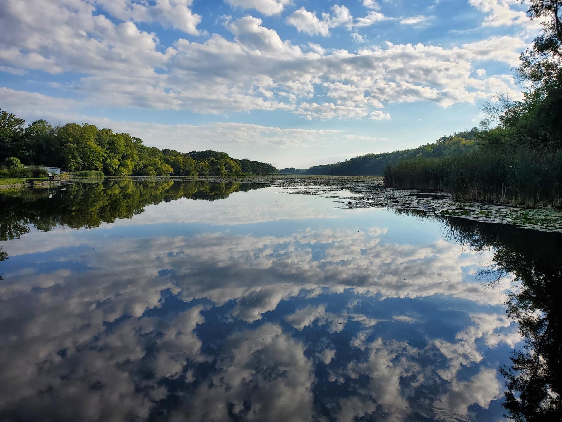 Wide lake reflecting dramatic white clouds and blue sky, surrounded by trees.