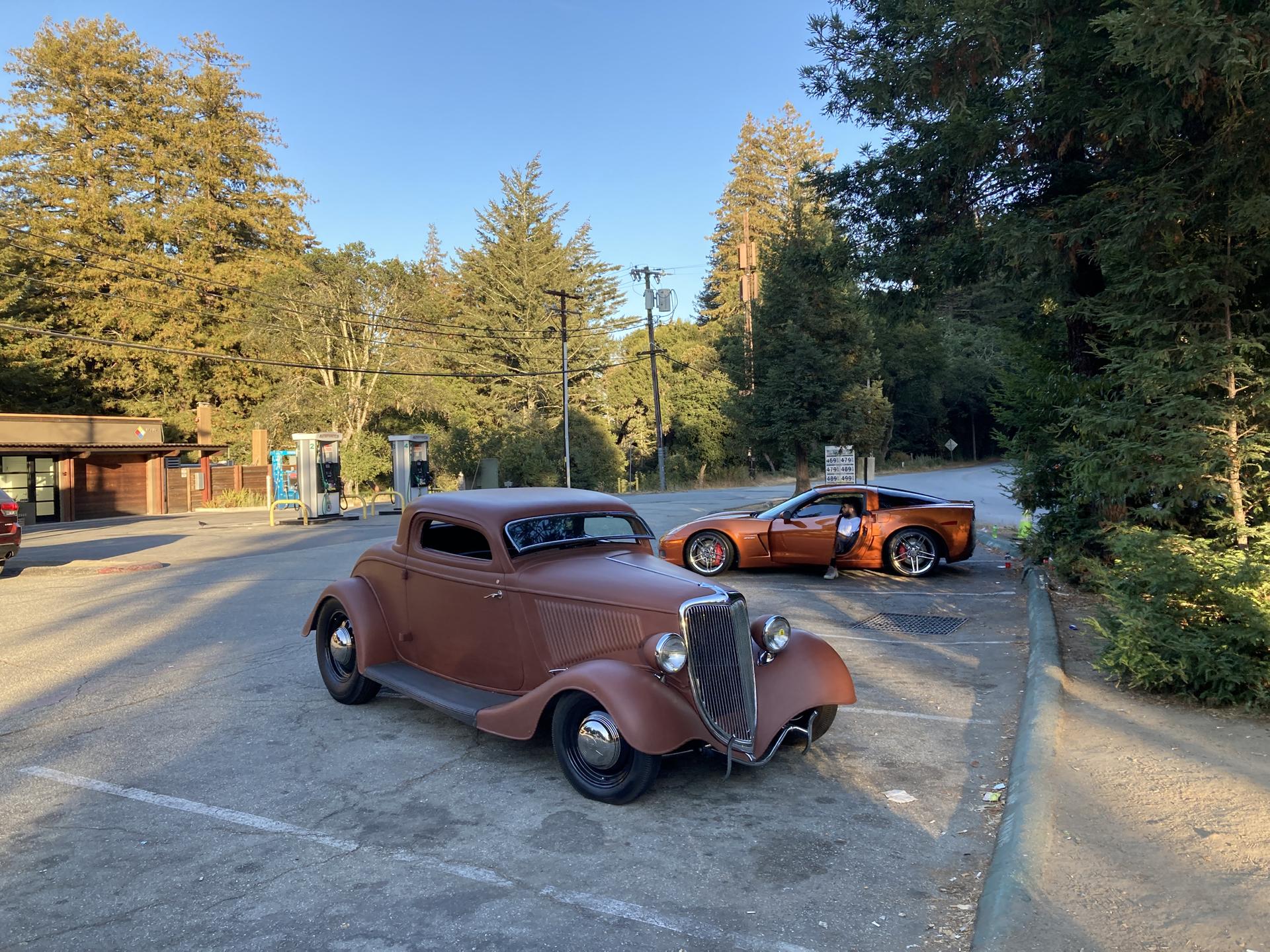 Two classic cars parked in a lot with trees and buildings.