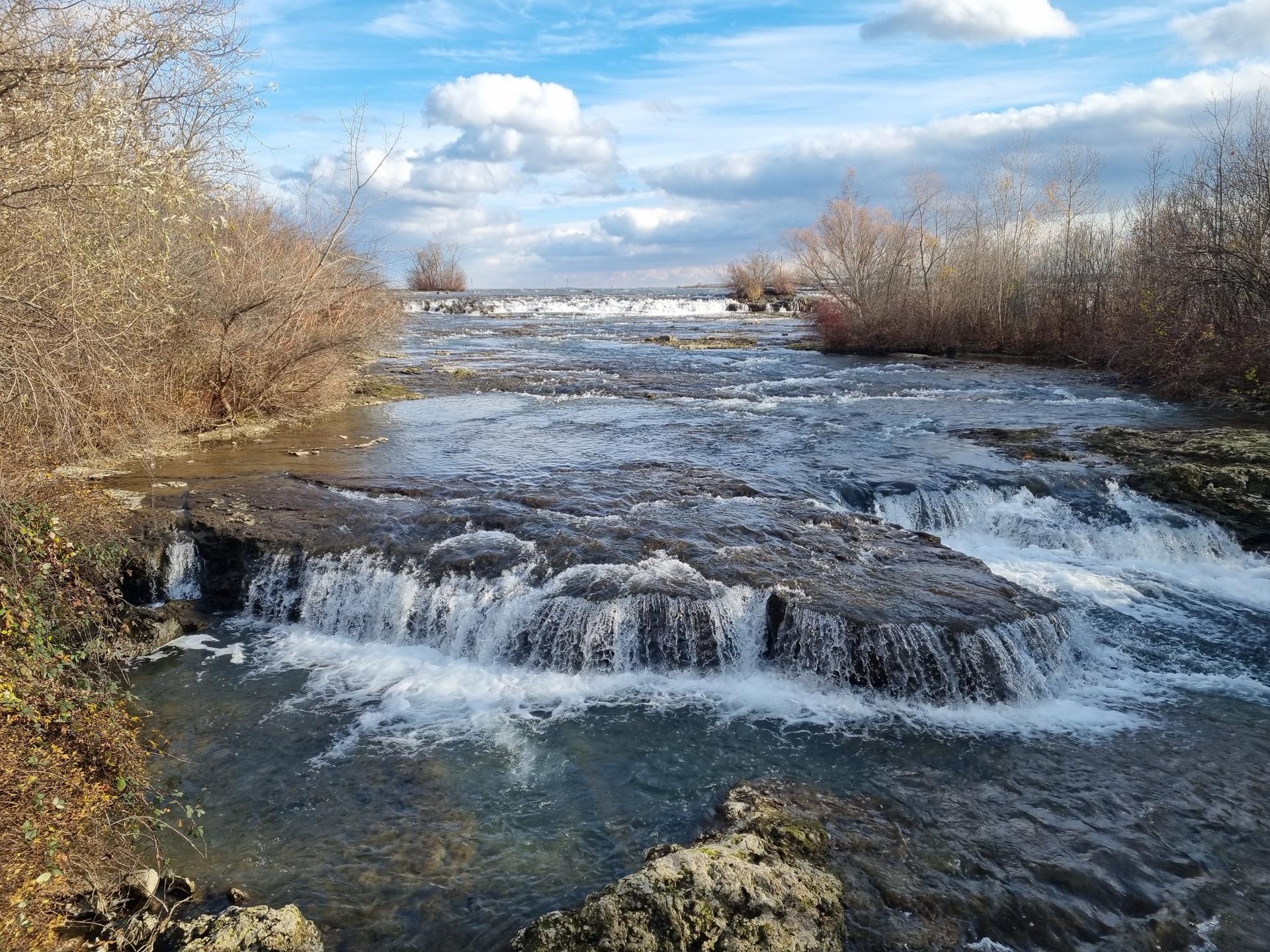 Wide river with small waterfalls, bare trees, and a cloudy blue sky.