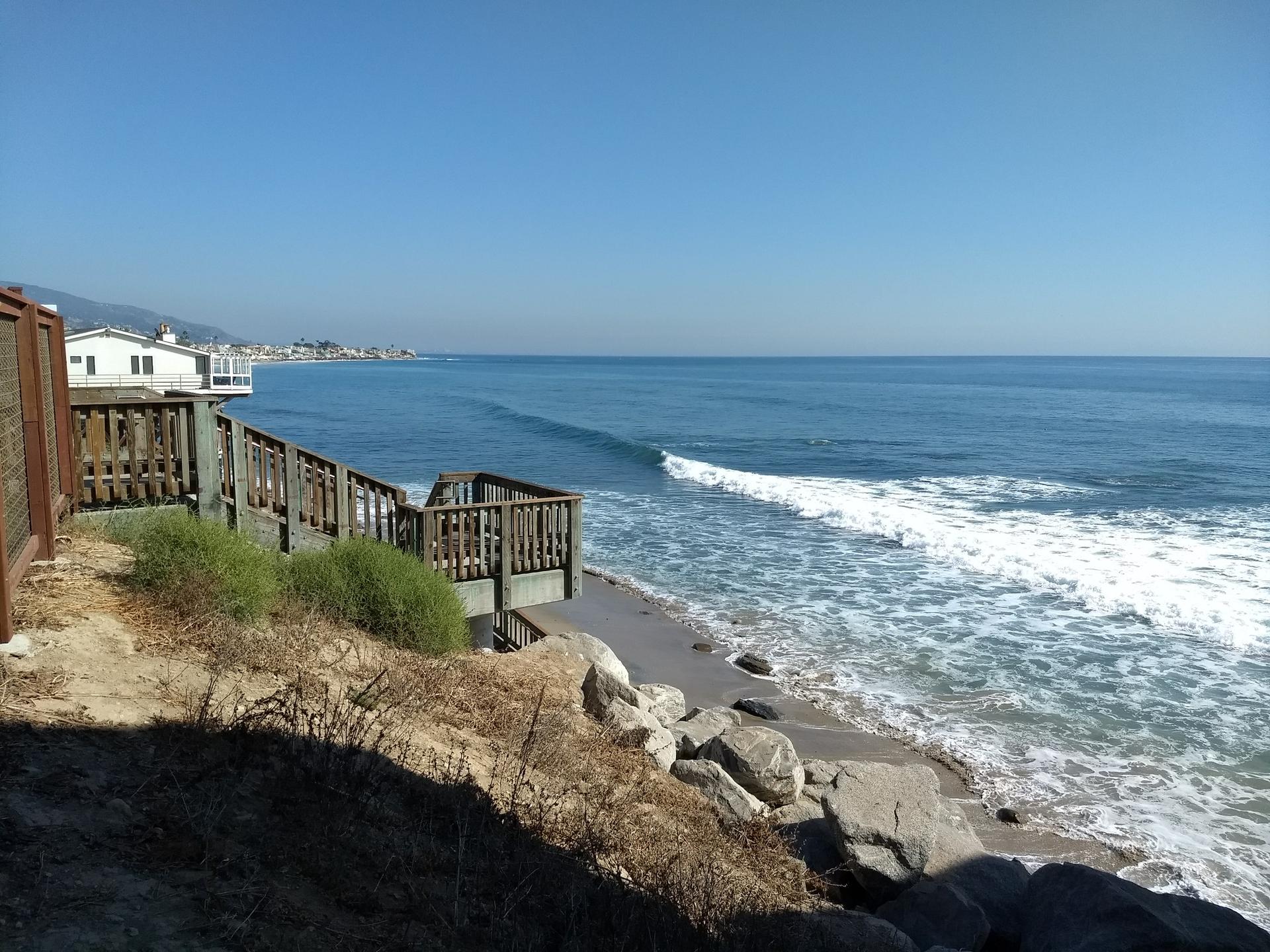 Wooden deck and stairs overlooking a sunny beach with crashing ocean waves.