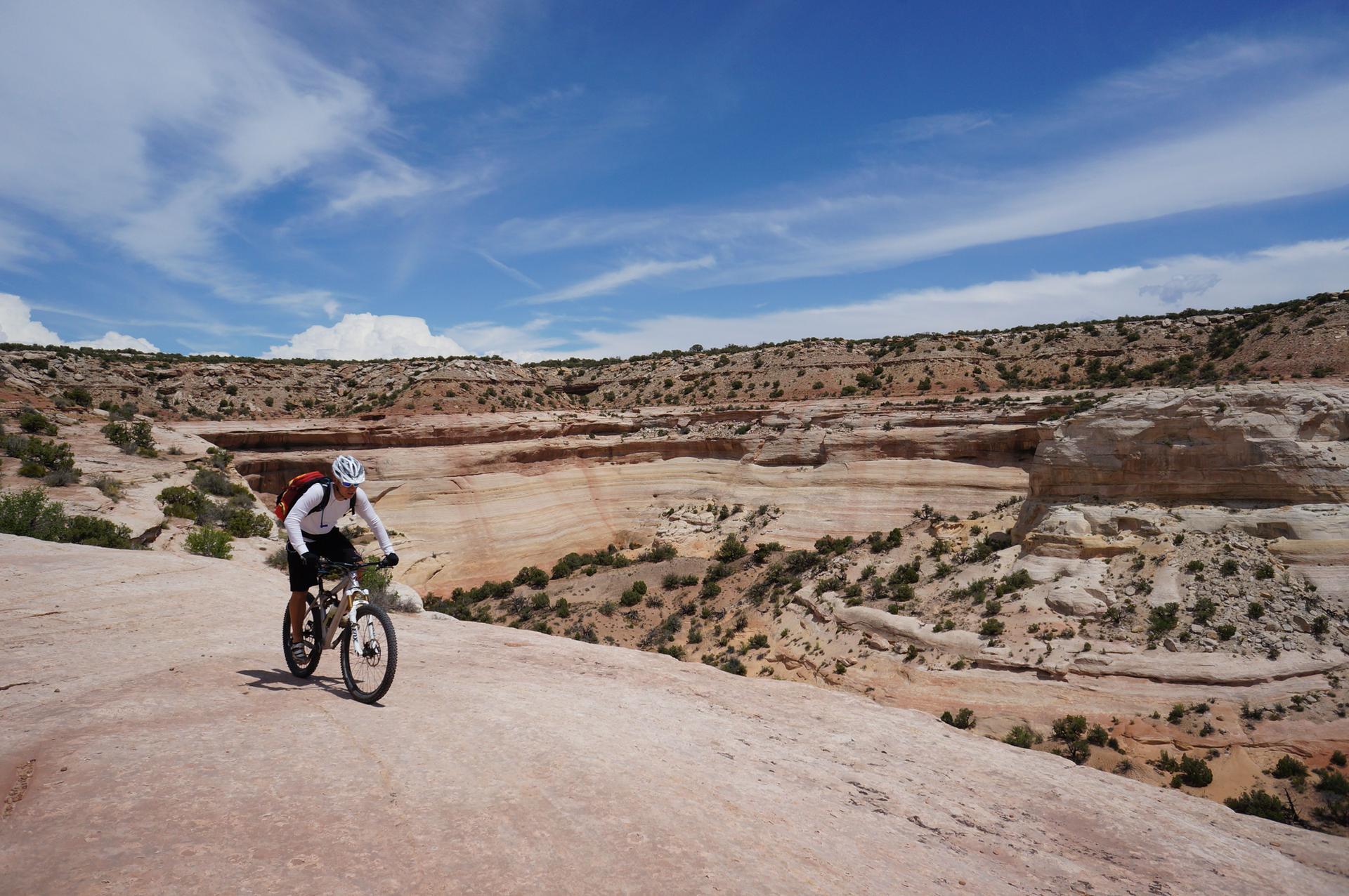 A mountain biker rides a rock slab overlooking a wide, sunny canyon.
