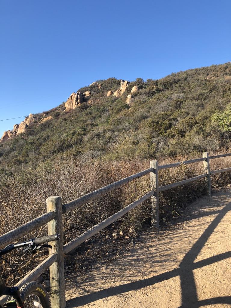 A dirt path with a wooden fence alongside a brushy hillside under a clear blue sky.
