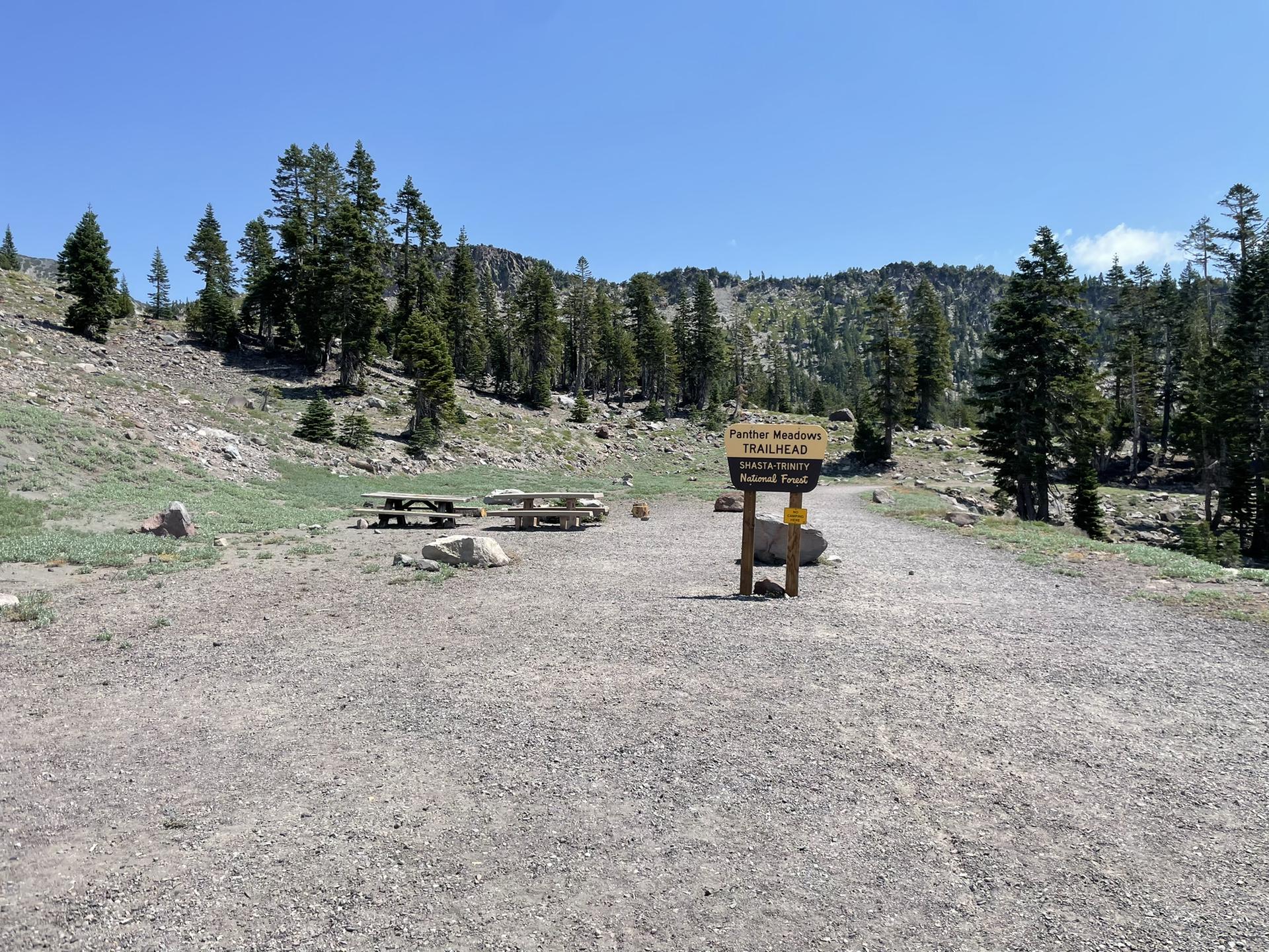 Gravel trailhead with picnic tables and a sign, surrounded by pine trees under bright sun.