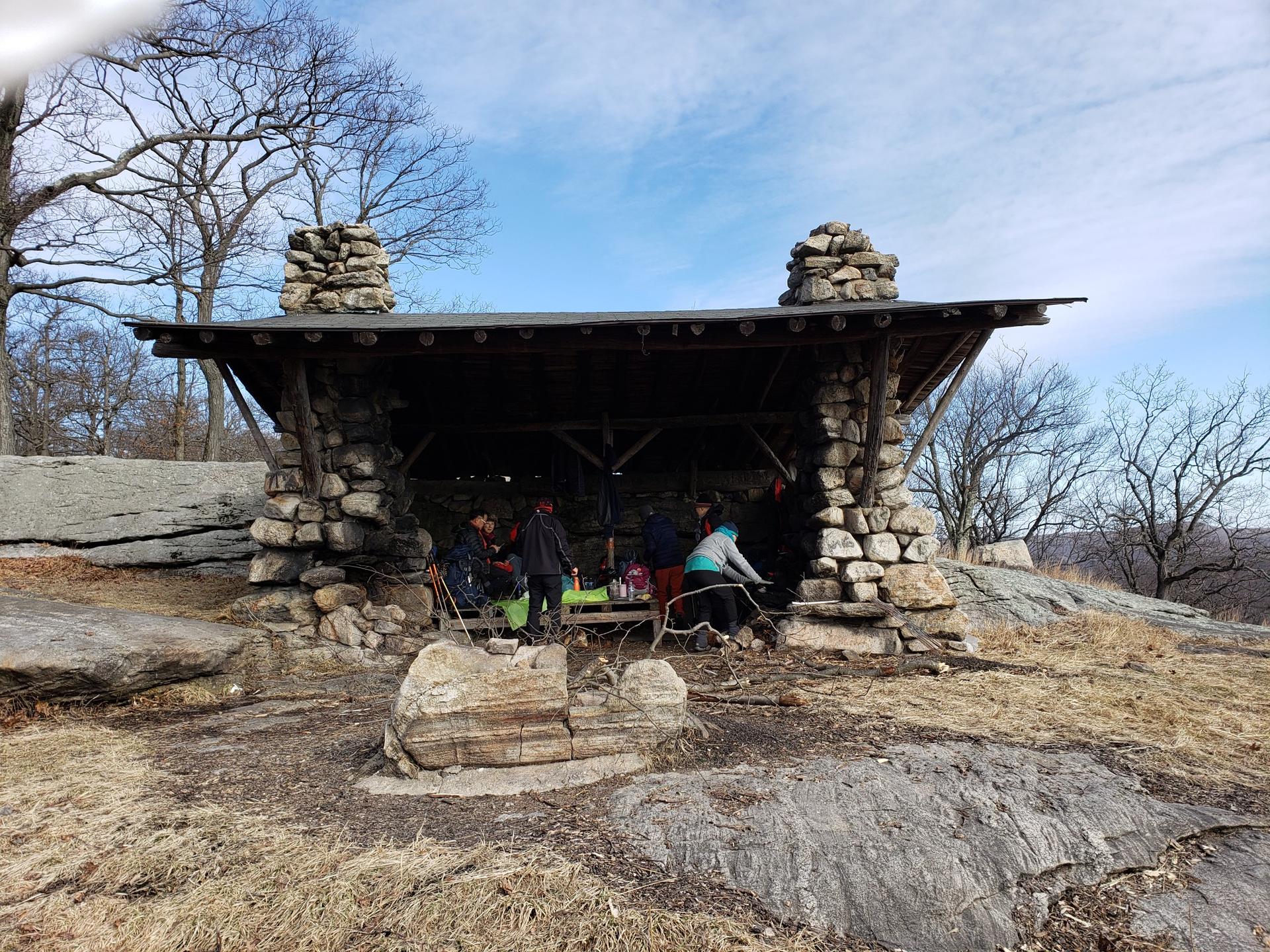 People gathered inside a rustic stone and wood shelter on a rocky hillside.