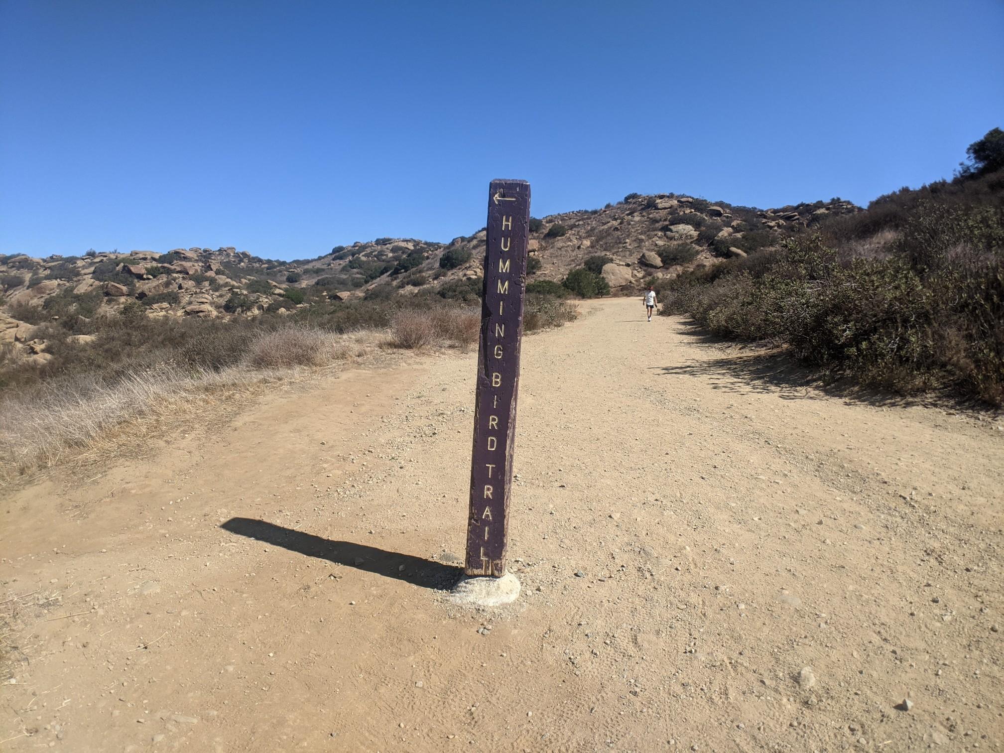 Trail sign on a dirt path, pointing left, with distant hills and a person.