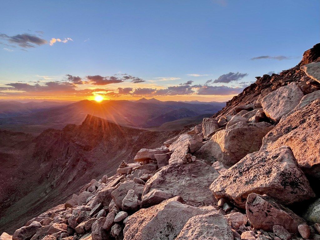 Golden sunset over a vast mountain range with large rocks in the foreground. - Mount Bierstadt Trail