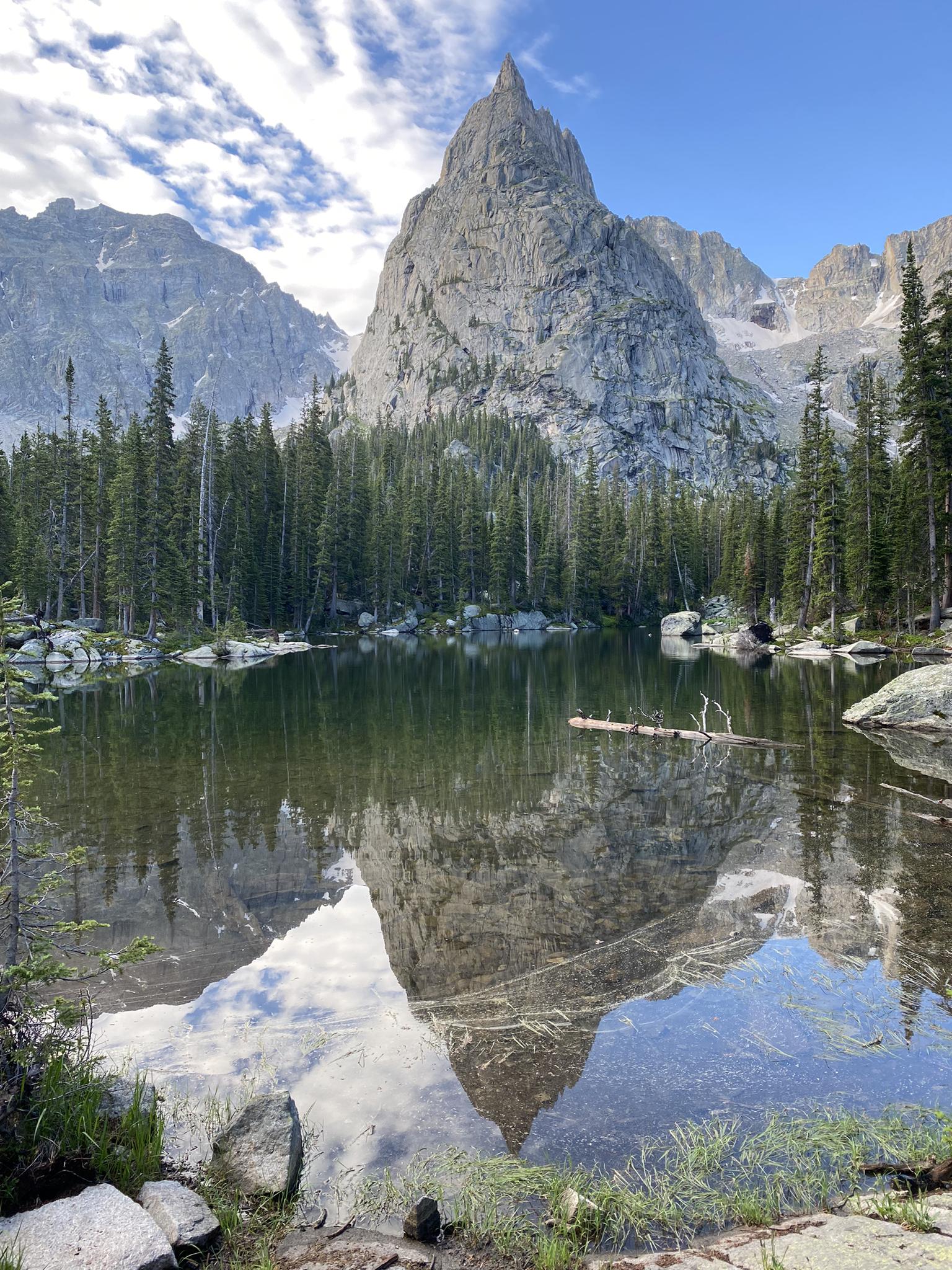 Jagged mountain peak reflected in a calm lake surrounded by pine trees on a clear day.