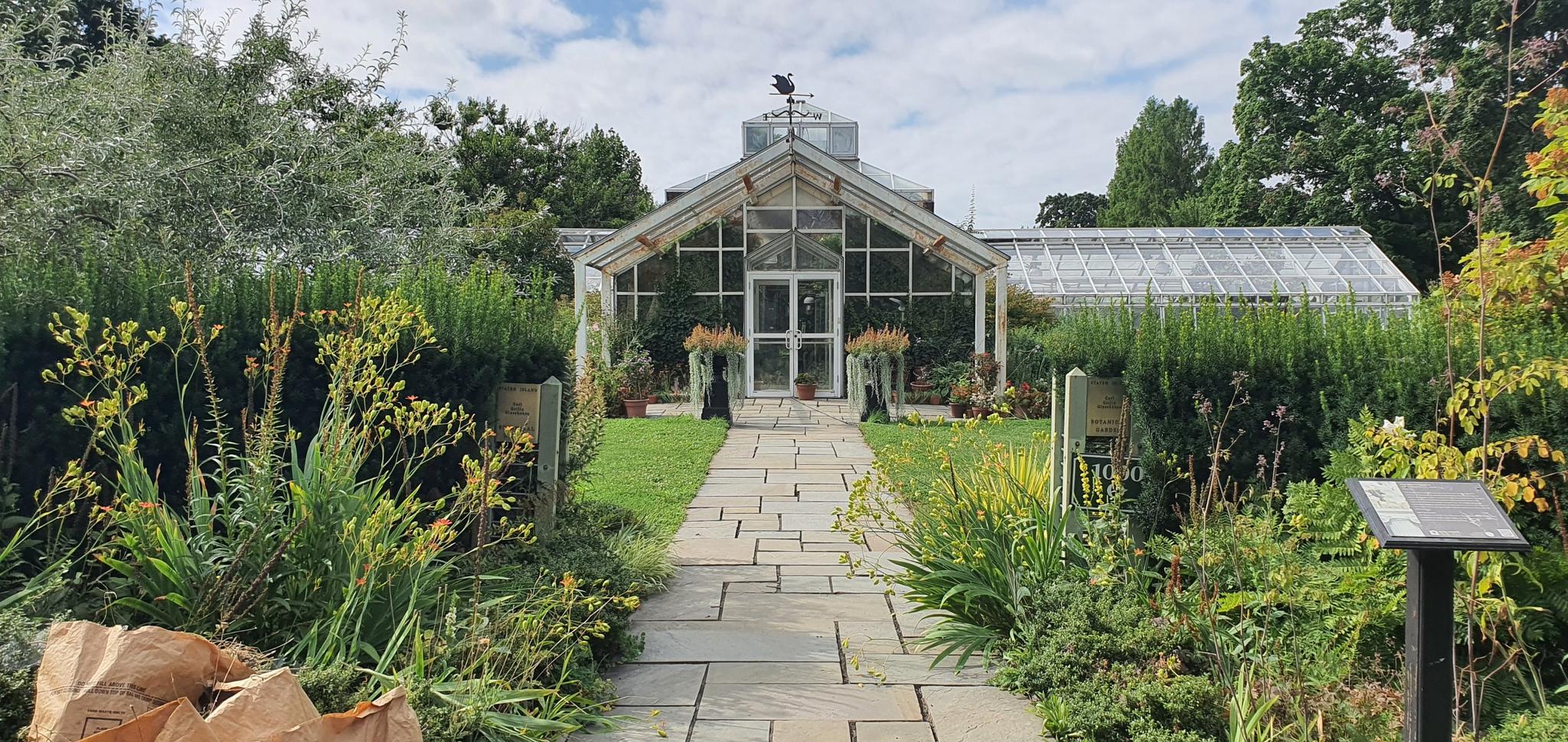A stone path leads to a large glass greenhouse surrounded by green plants.