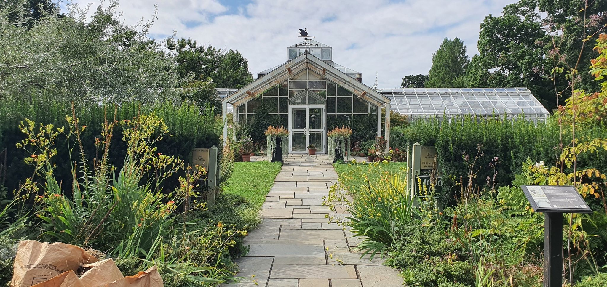 A stone path leads to a large glass greenhouse surrounded by green plants. - Staten Island loop