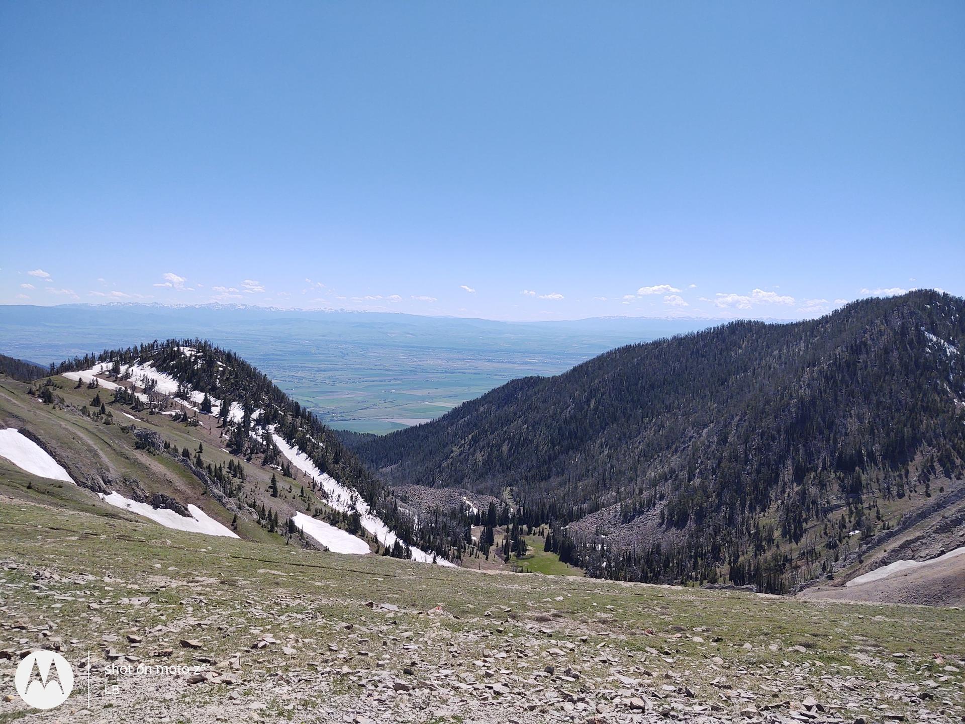 Rolling forested mountains with snow patches, overlooking a wide valley and plains.