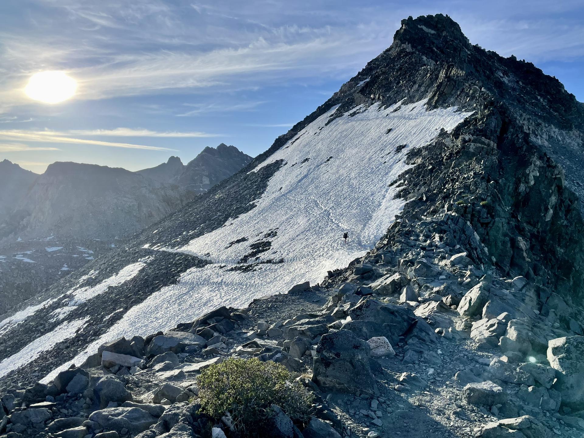 A person hikes up a steep, snowy mountain slope under a bright sun.