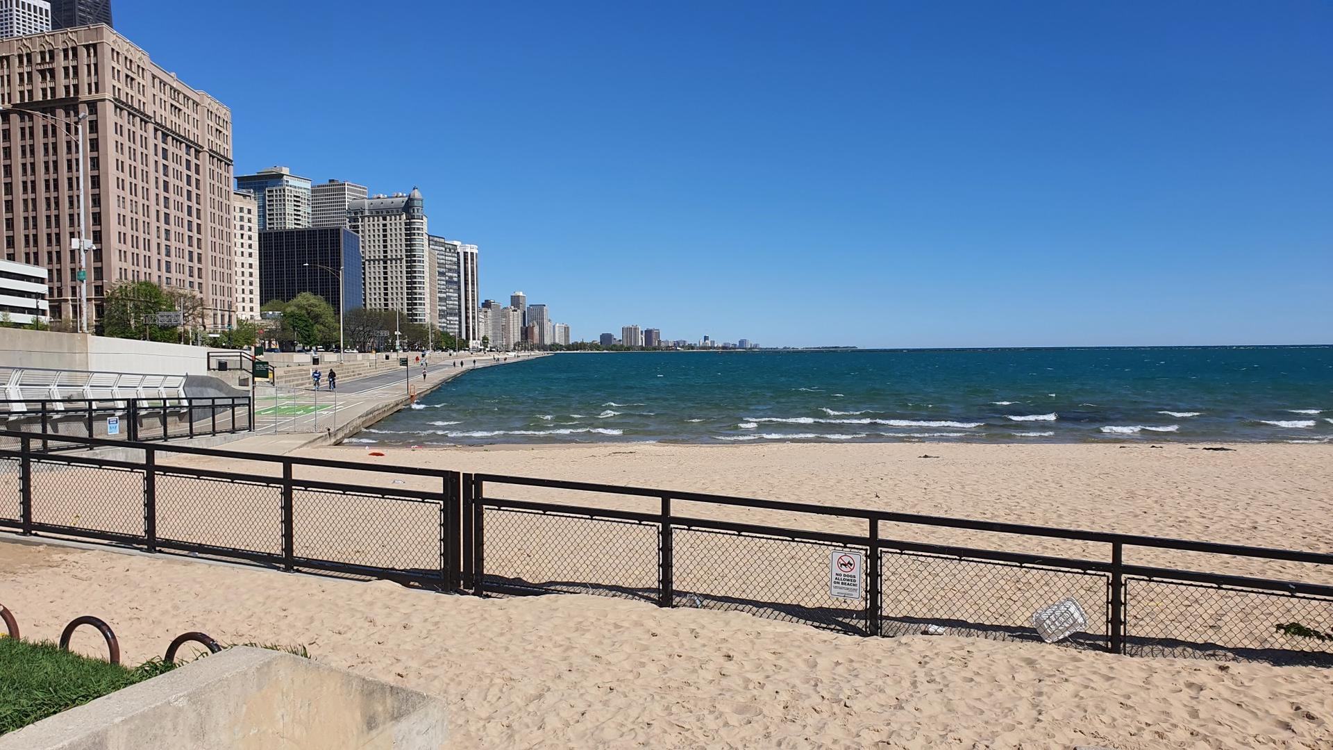 Sandy beach with a fence, city skyline, and blue water under clear sky.