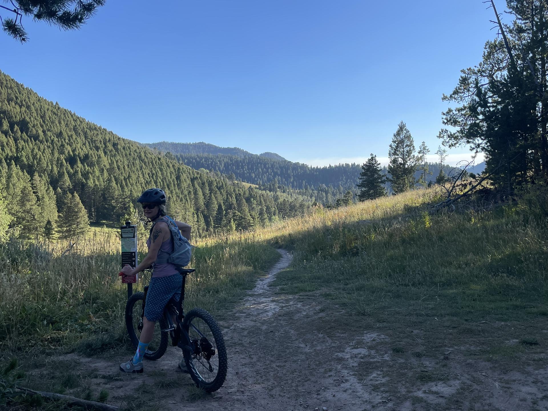 A person with a mountain bike stands on a dirt trail in sunny mountains.