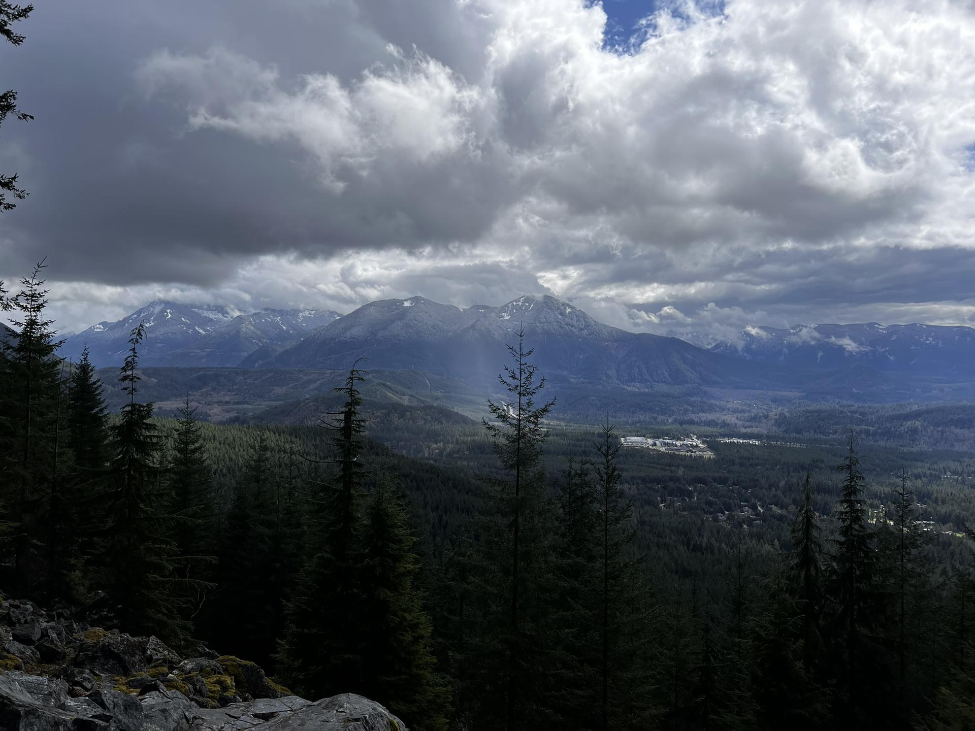 Dark evergreen forest with sun rays illuminating distant mountains under a cloudy sky.