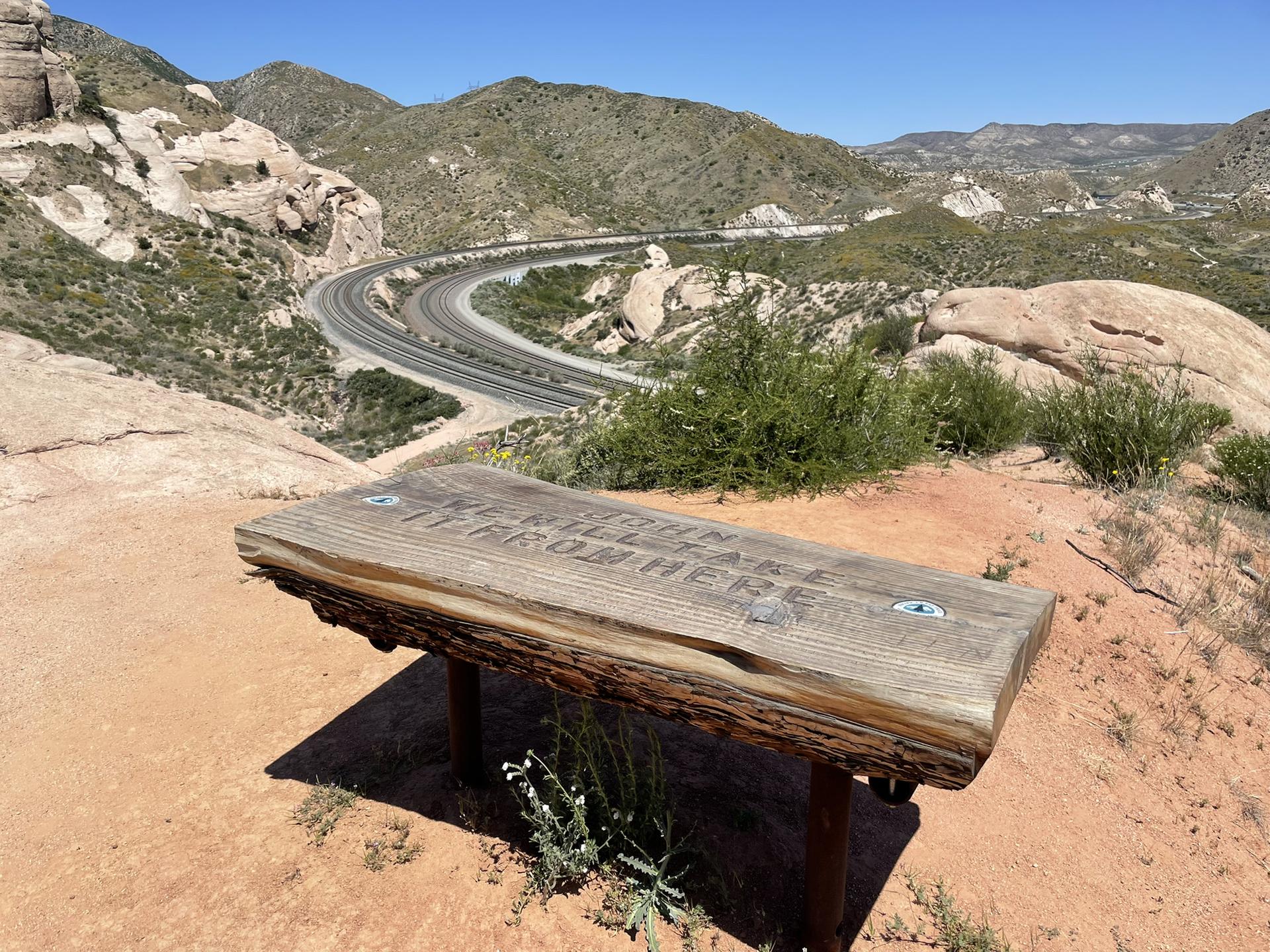 A rustic wooden bench overlooks a winding road through arid hills.