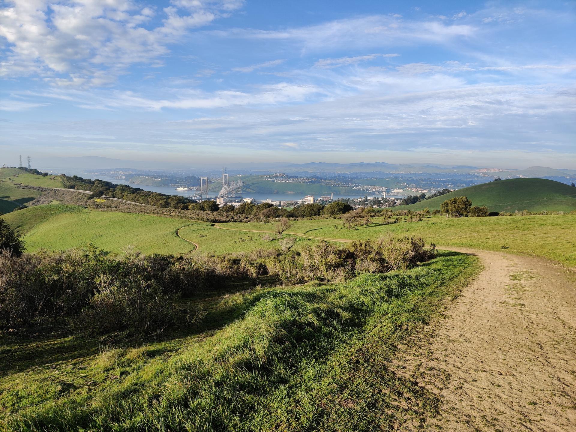 Dirt path winding through green hills with a distant city view.