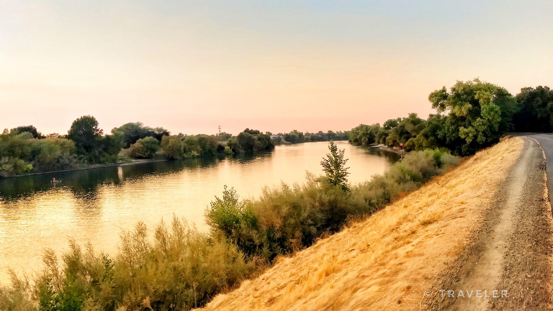 Golden sunset light on a river, with a dirt path and trees along the bank.