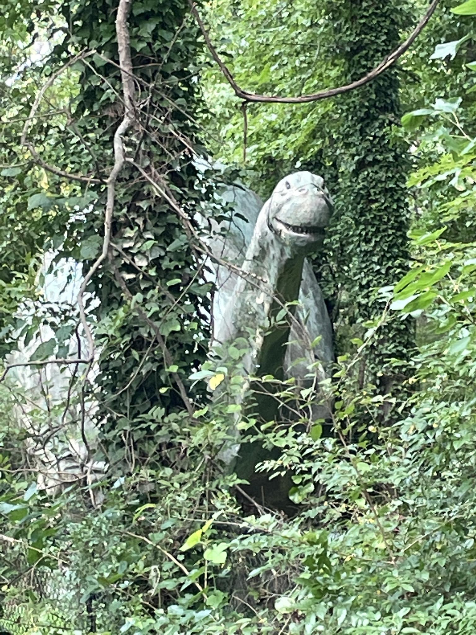 A large dinosaur statue is partially hidden by thick green foliage.