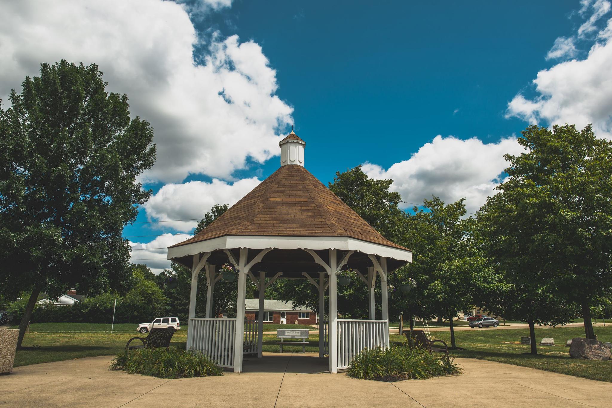 White gazebo with a brown roof under a bright blue sky and green trees.