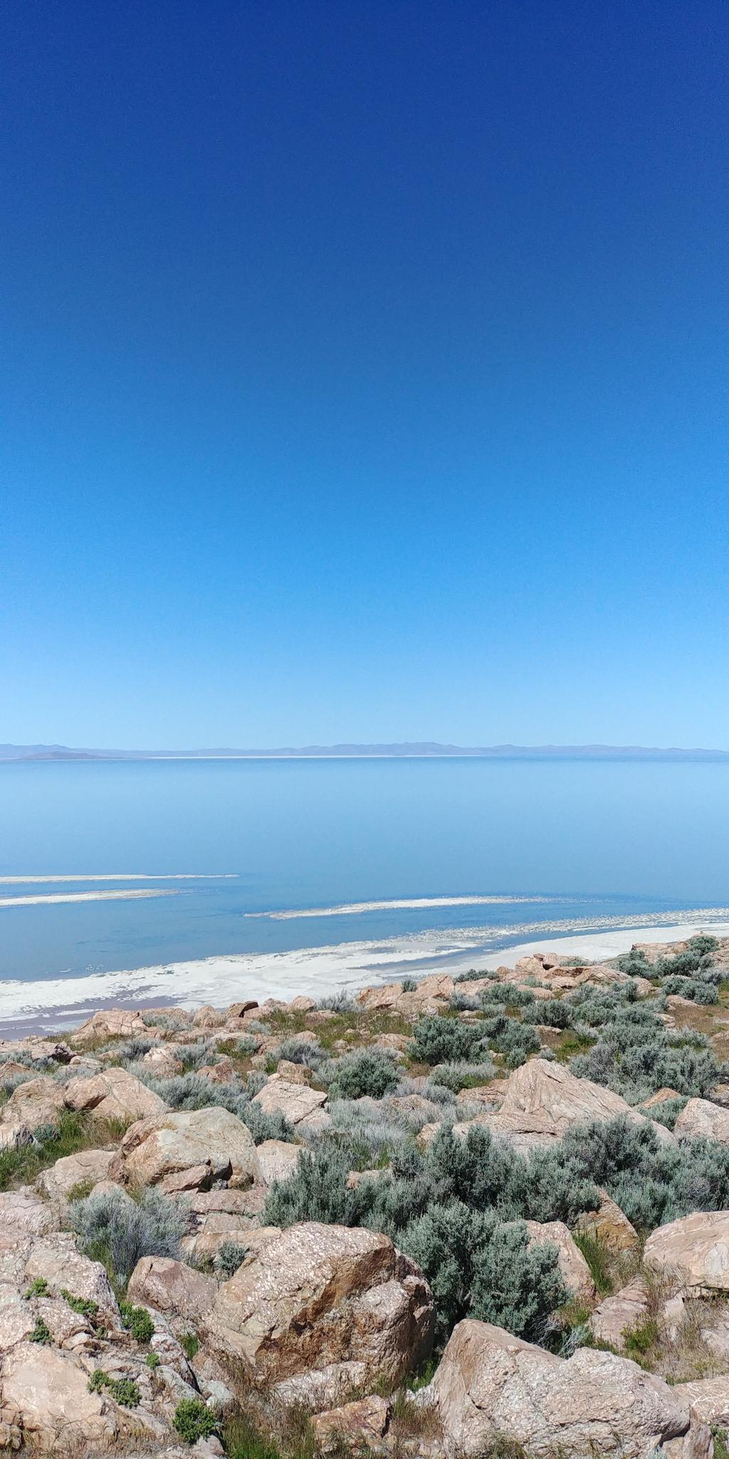 Rocky foreground with bushes overlooking a vast, calm lake under a clear blue sky.