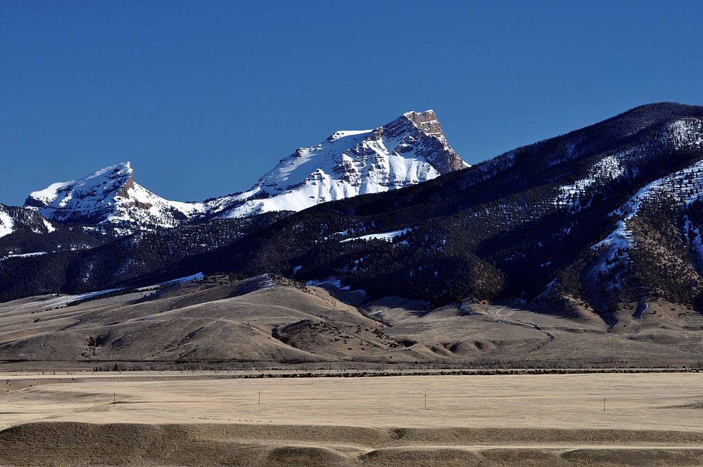 Majestic snow-capped mountains overlook a wide, dry field under a clear blue sky.