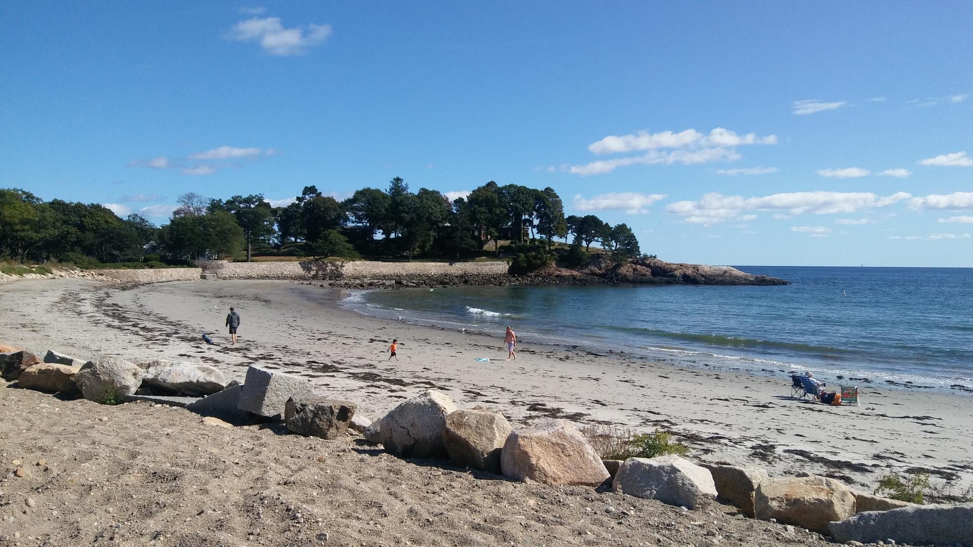 Sandy beach with scattered rocks, ocean waves, and distant people under blue sky.