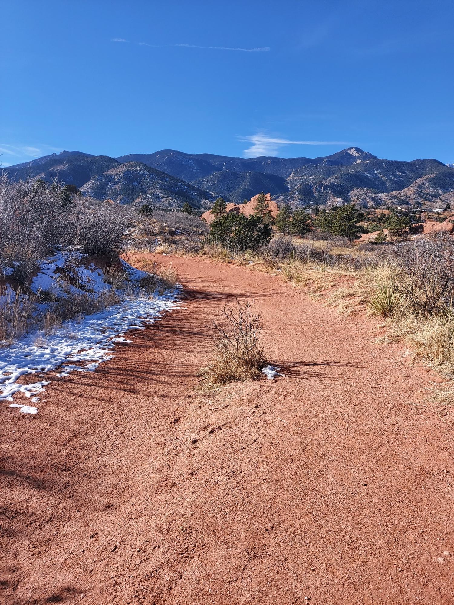 Red dirt path with snow patches leading to distant mountains.
