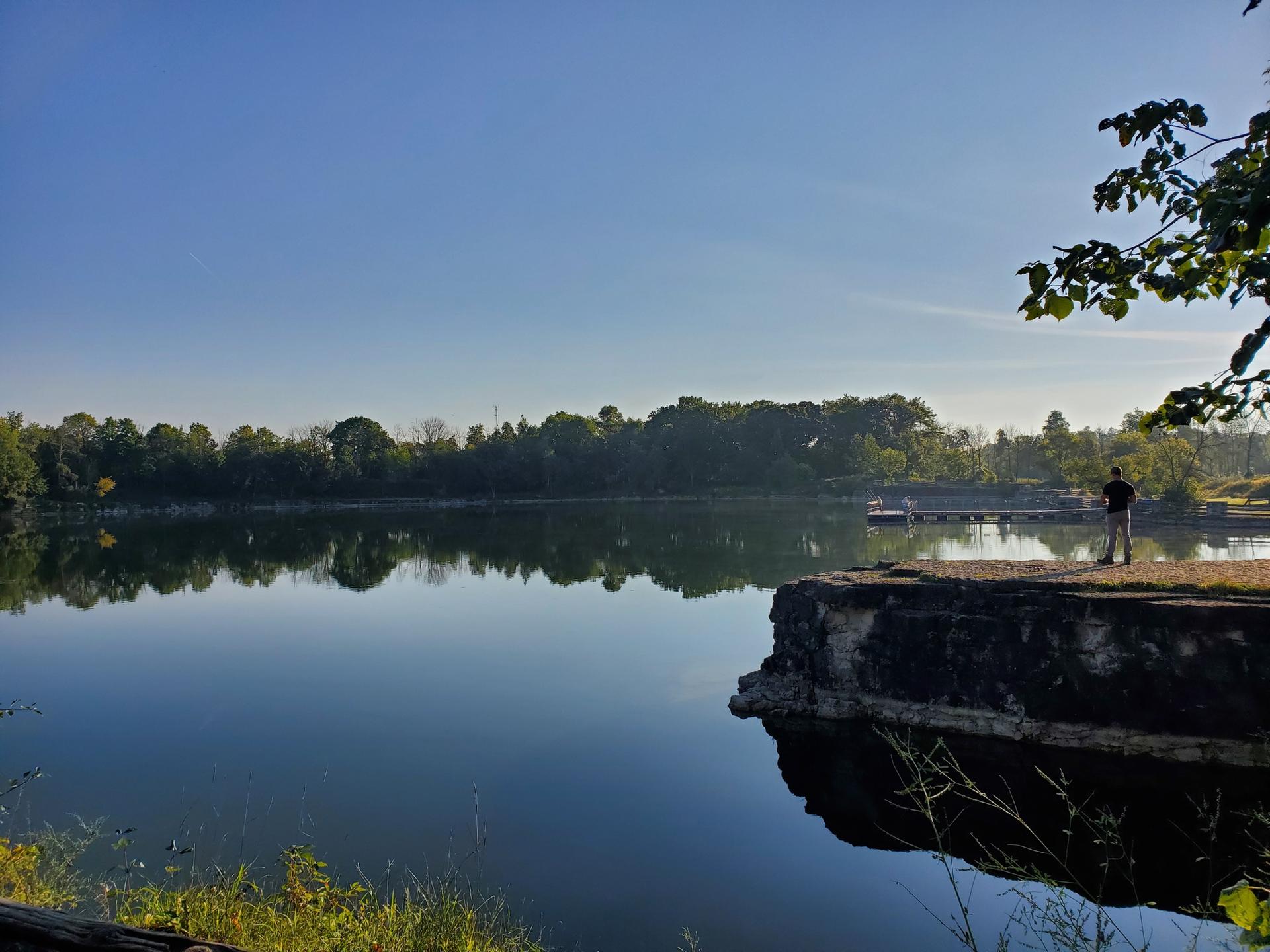 Calm lake reflecting trees, with a person standing on a stone pier.
