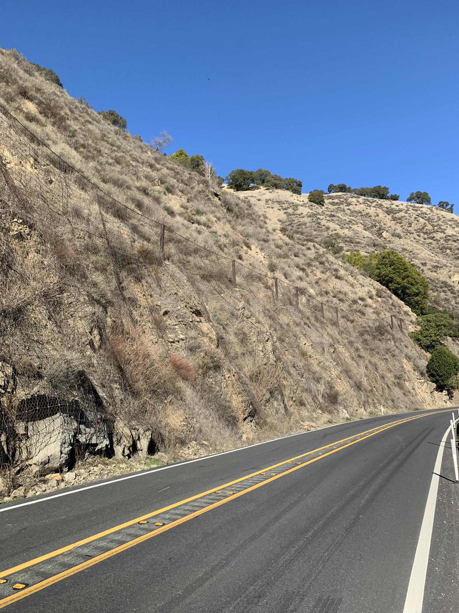 A paved road alongside a steep, dry, rocky hillside under clear blue sky.