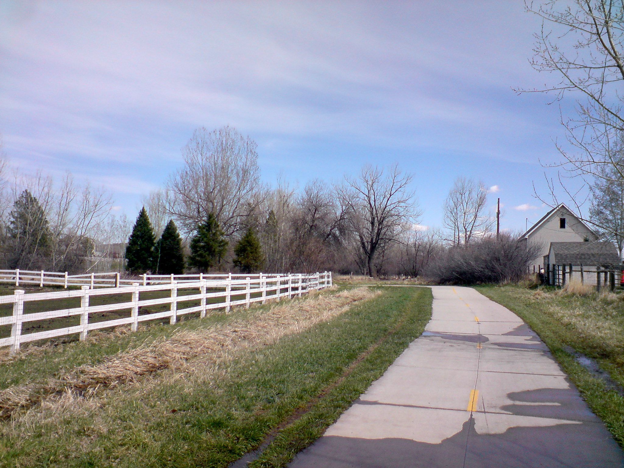 Paved path alongside a white fence, bare trees, and a distant building. - Spring Creek, Fossil Creek, and Power Trail Loop