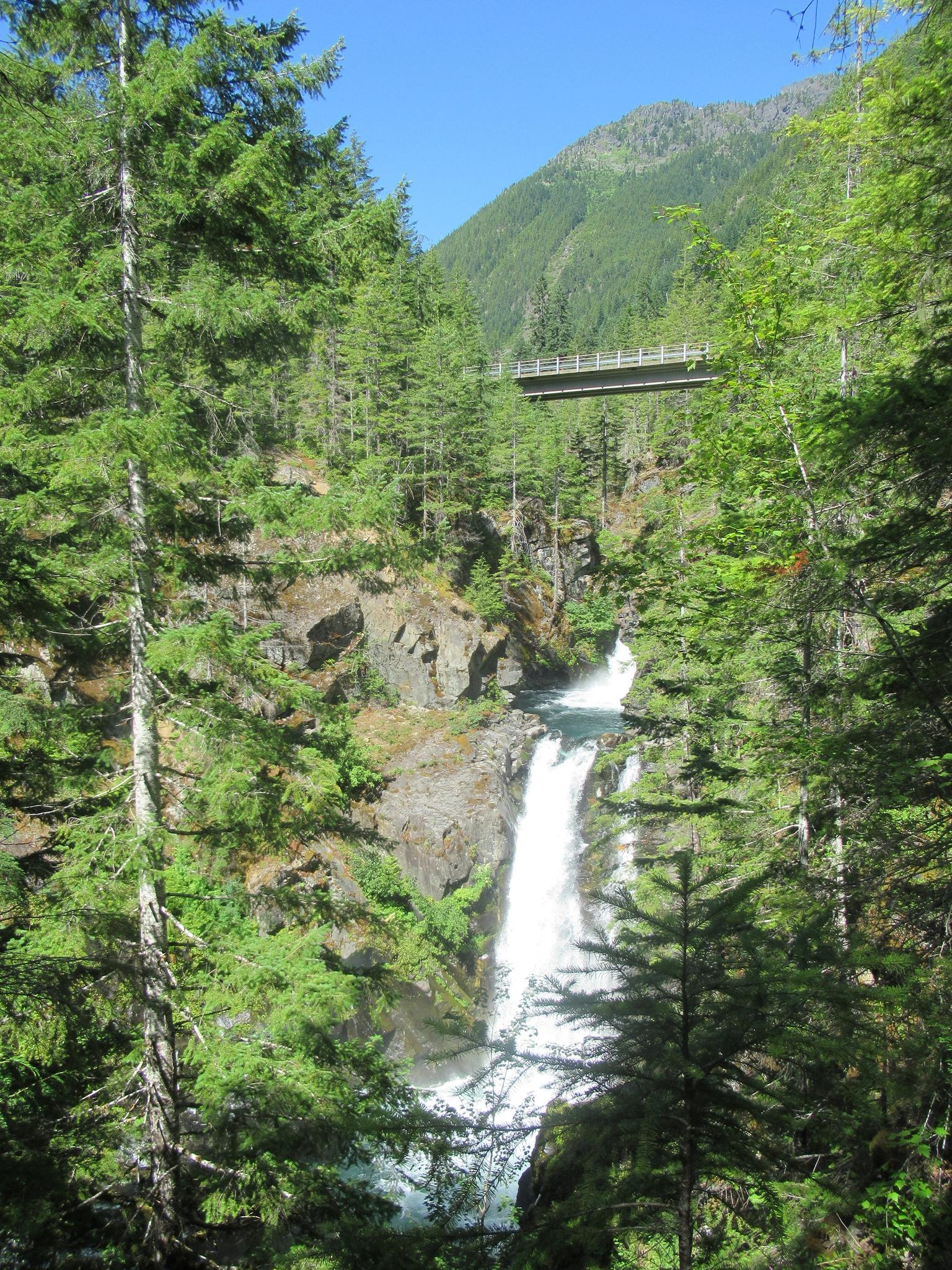 Waterfall cascades through a lush forest gorge under a distant bridge, sunny day.