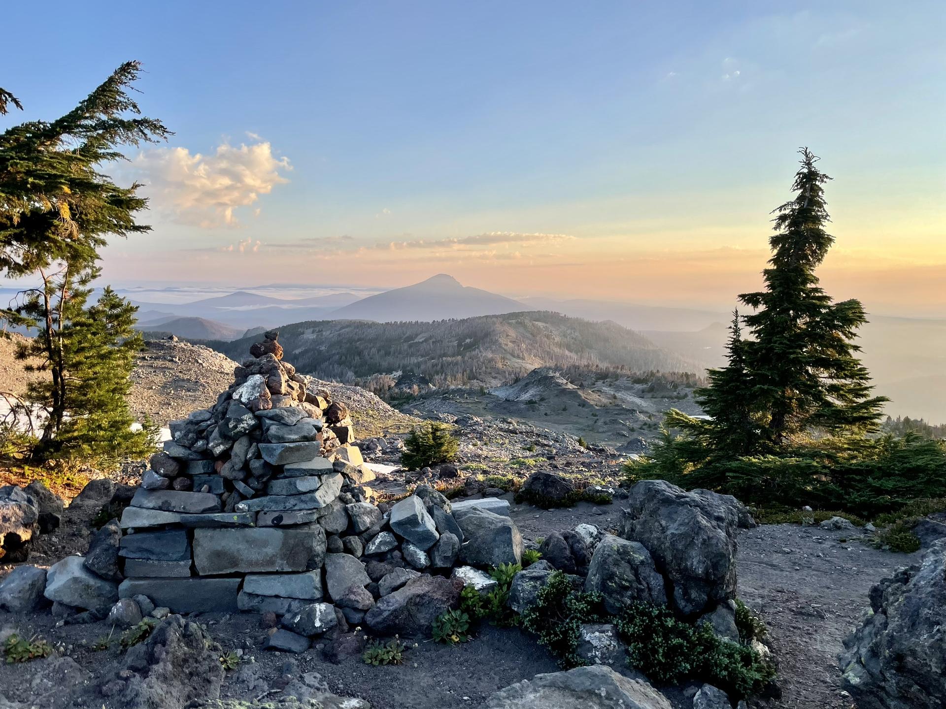 Stone cairn on a mountain peak overlooking hazy valleys at sunset.