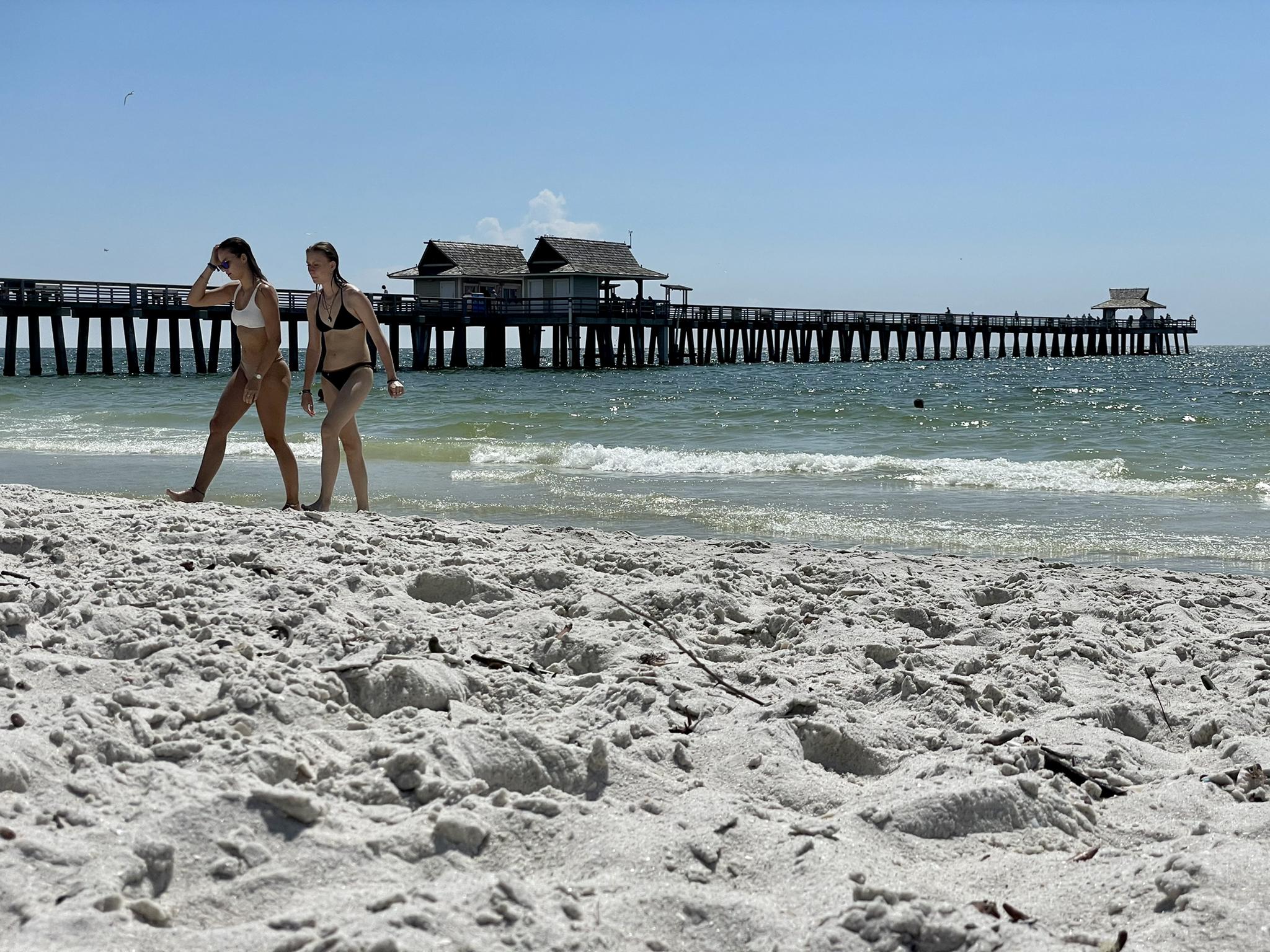 Two women walk on a sunny beach with a long pier.