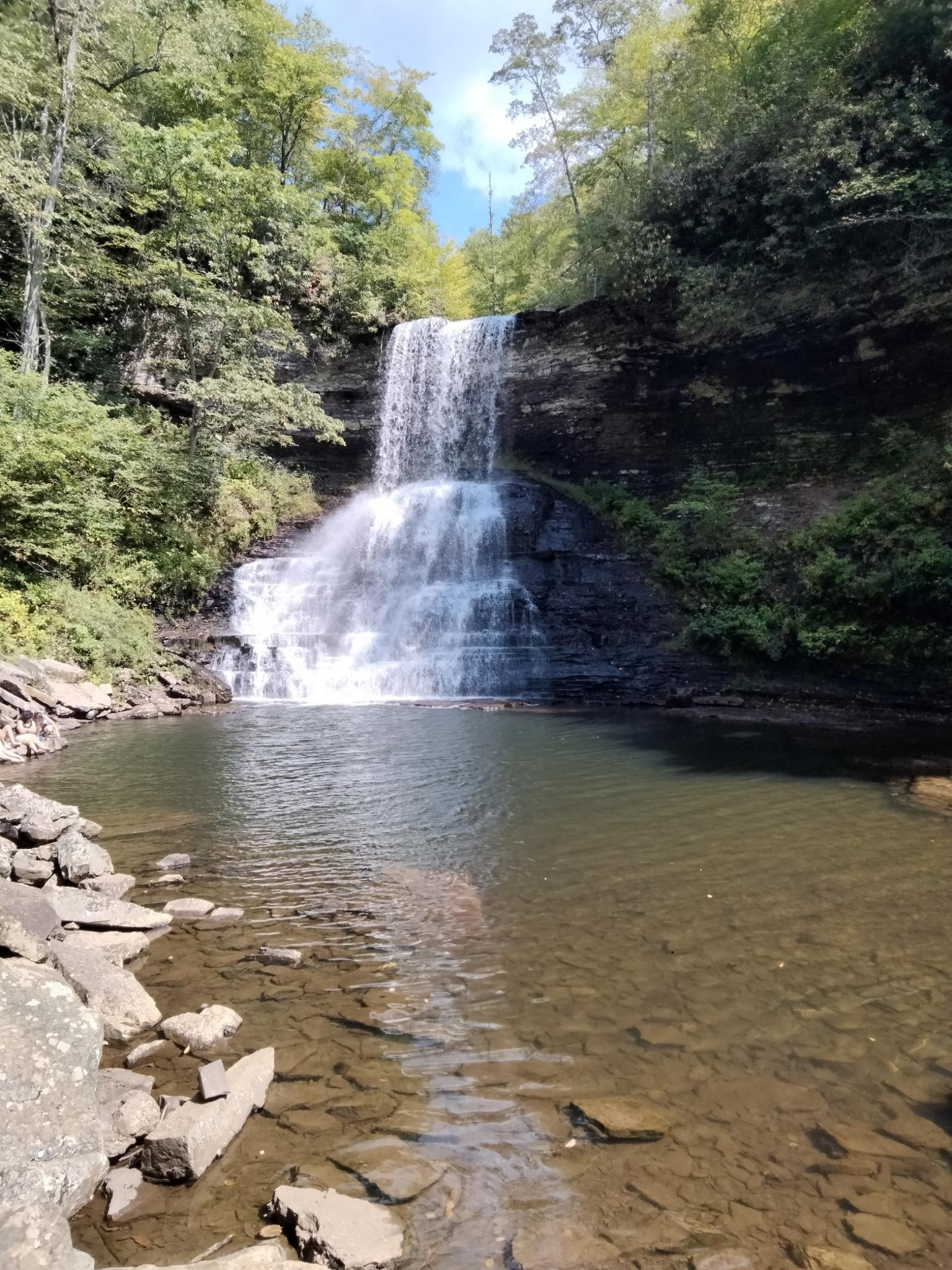 Waterfall cascading into a clear pool, surrounded by green trees under bright sky.