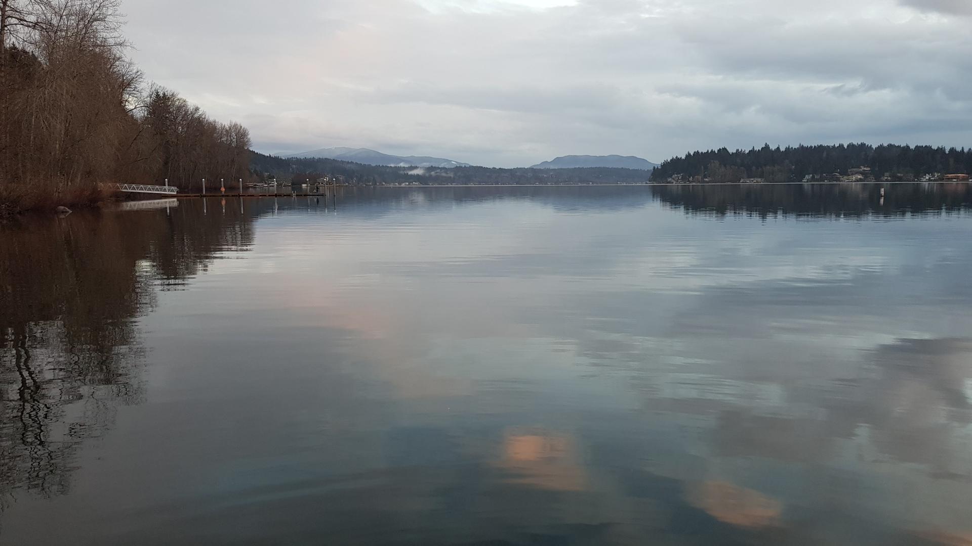Calm lake reflecting a cloudy sky, with forested shores and distant mountains.
