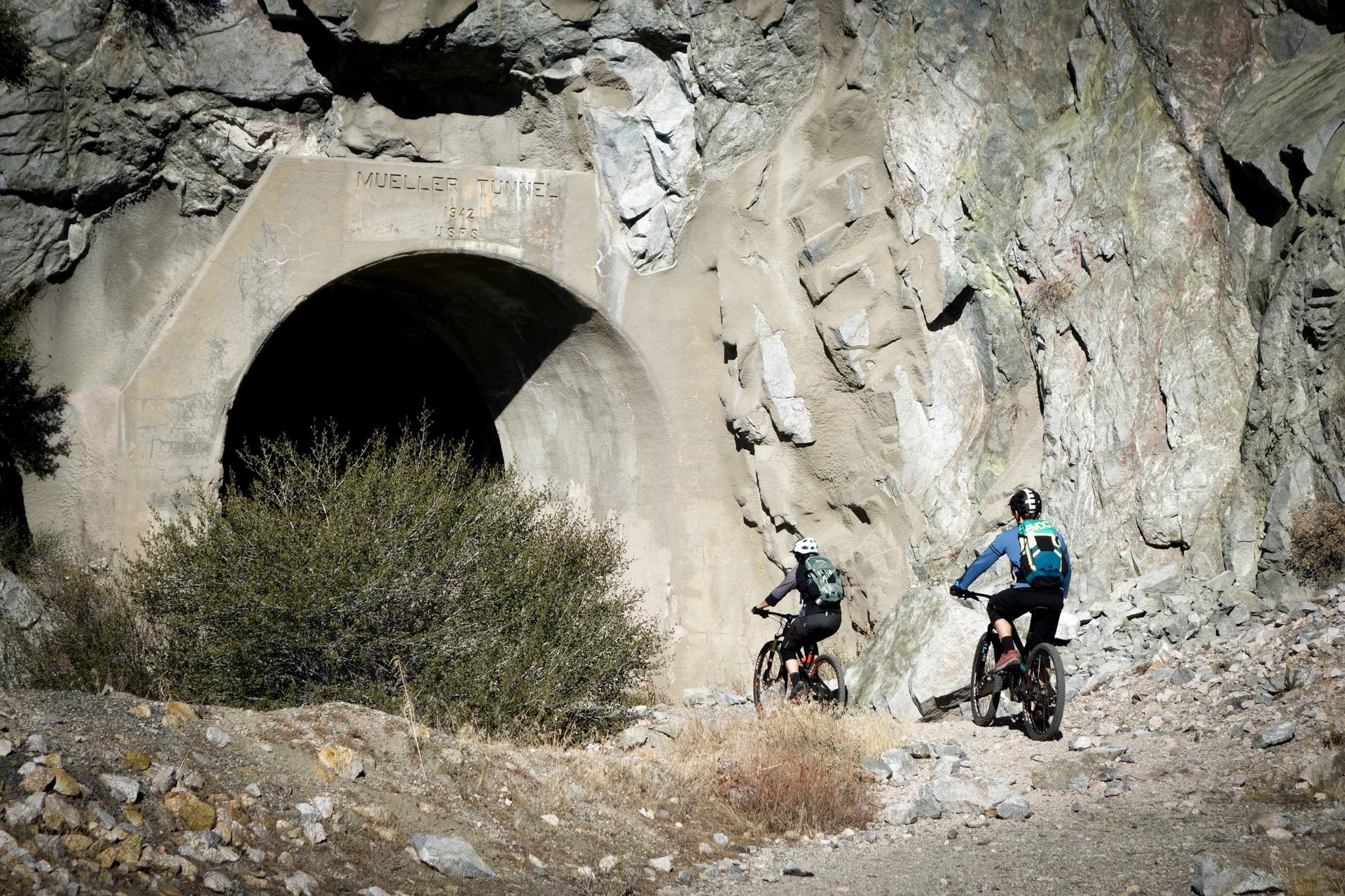 Two mountain bikers approach a dark tunnel entrance in a rocky landscape.