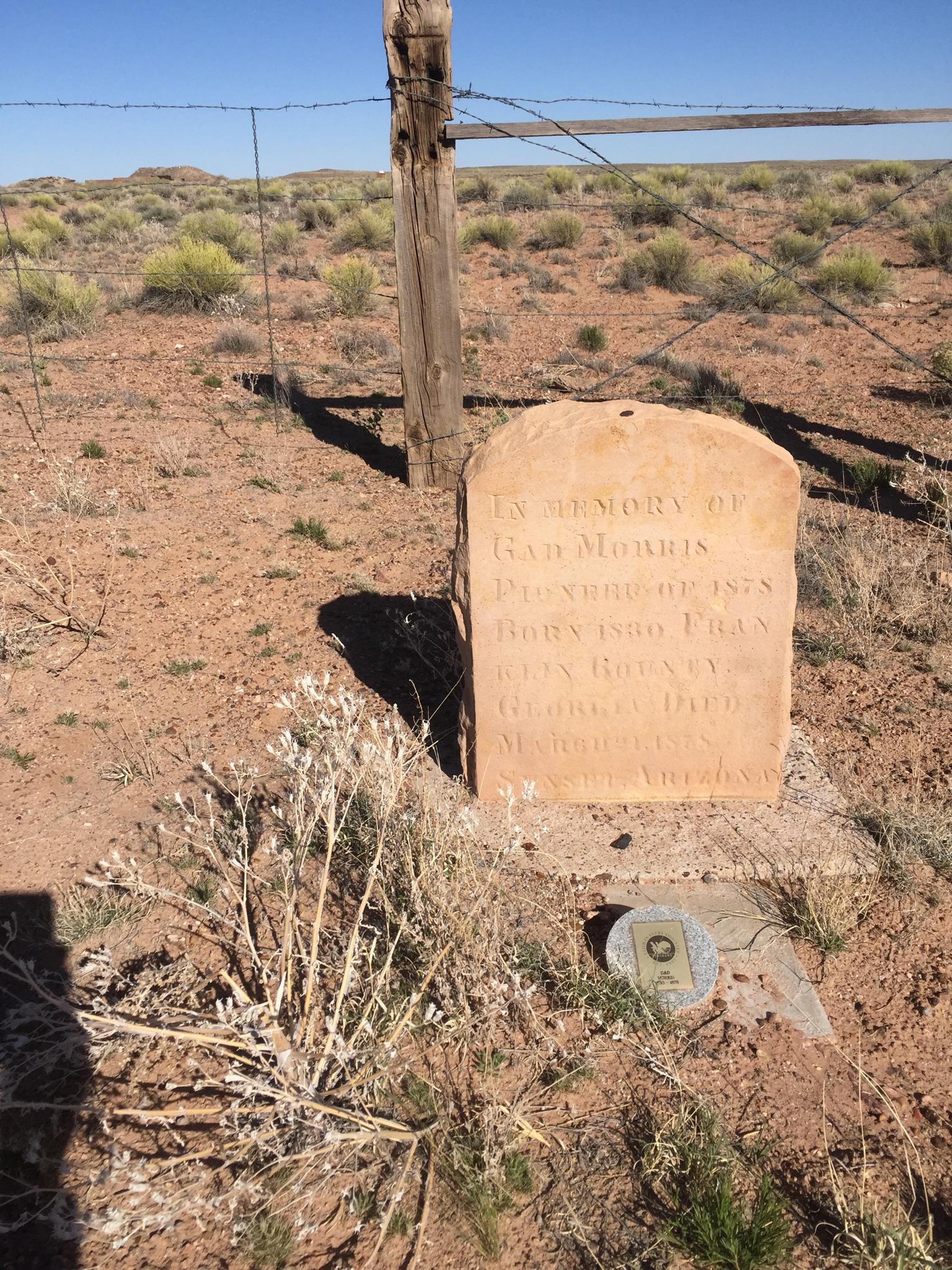 An old weathered stone headstone stands in dry, sparse landscape with a fence.