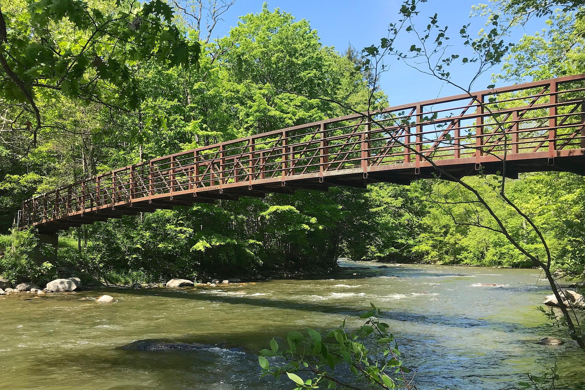 A long metal pedestrian bridge crosses a wide river under bright sun.