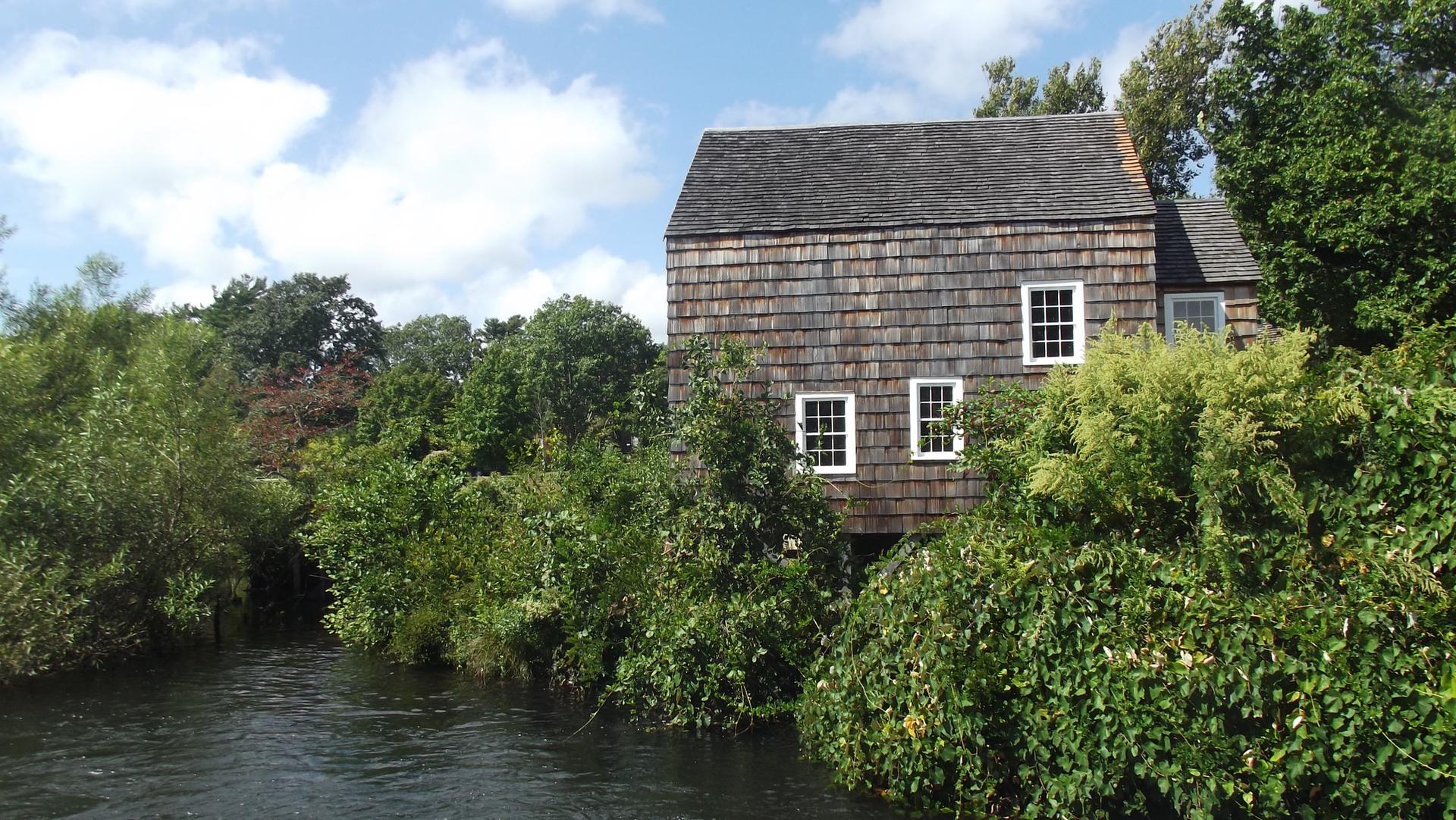 A rustic wooden building stands beside a river surrounded by lush greenery.