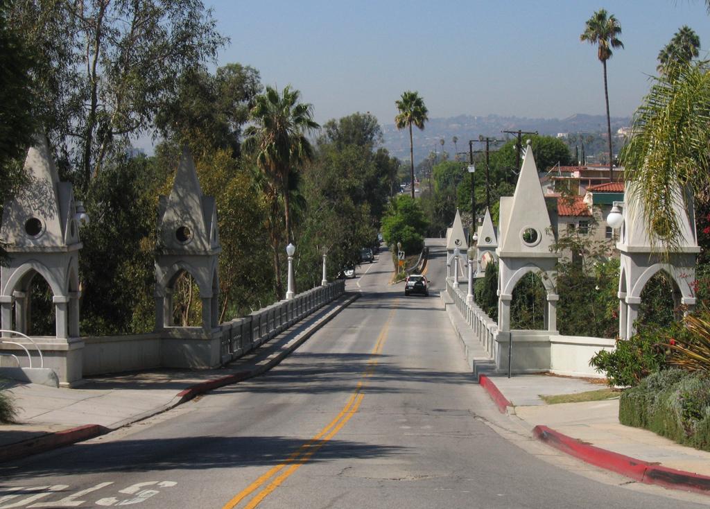 A sunny road over a bridge with decorative stone towers, surrounded by trees.