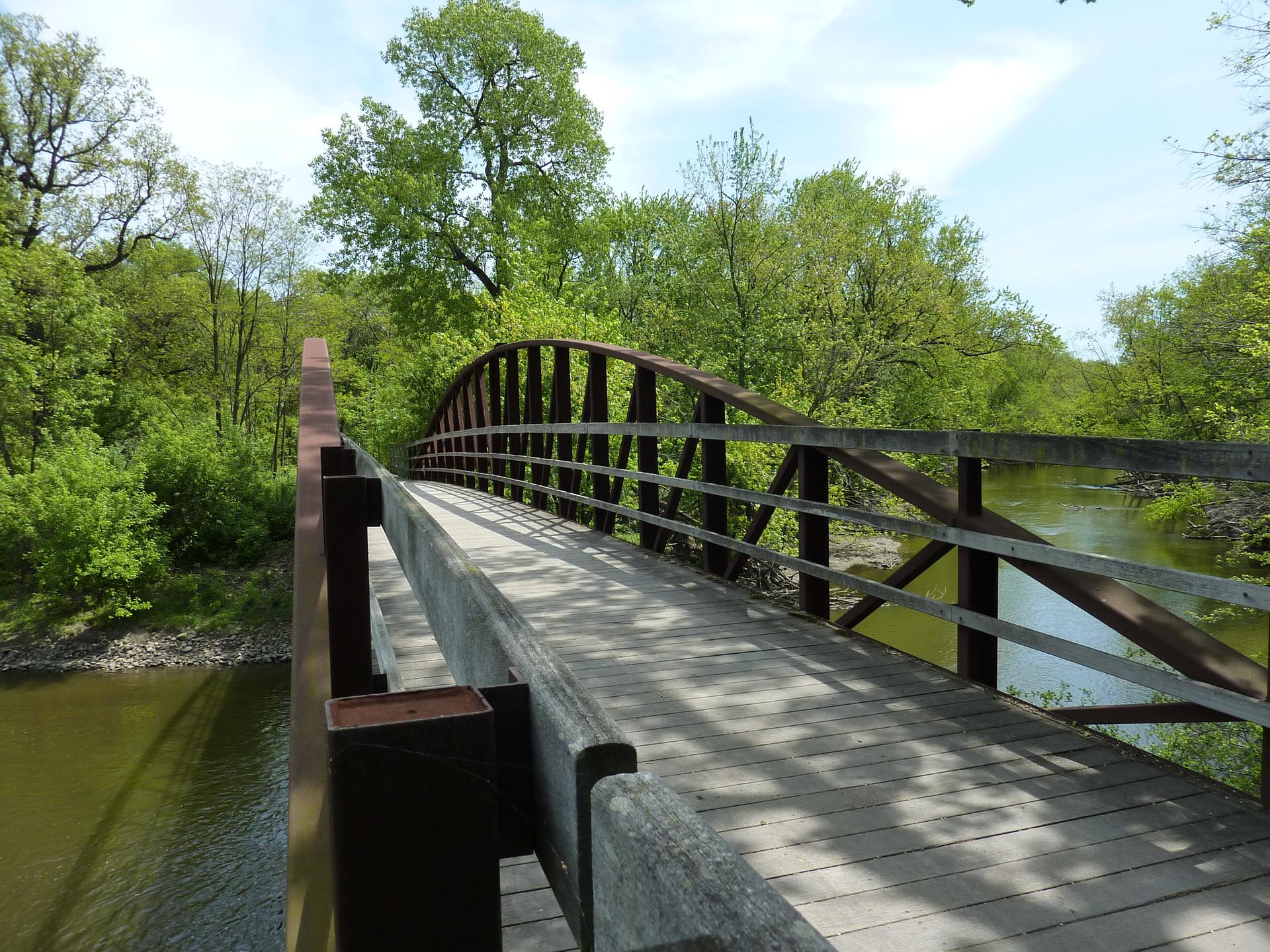 A wooden and metal arched bridge spans a river surrounded by green trees.