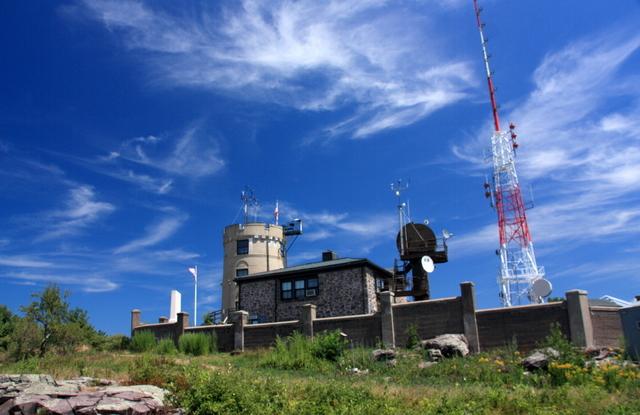 Stone building and tall communication towers on a sunny hill under a blue sky.