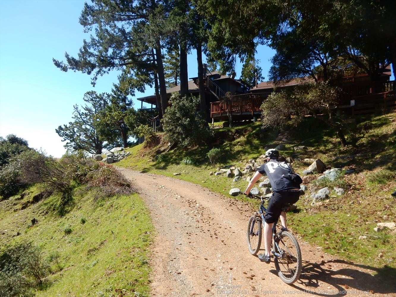 A person mountain biking up a sunny dirt road past trees and a building.