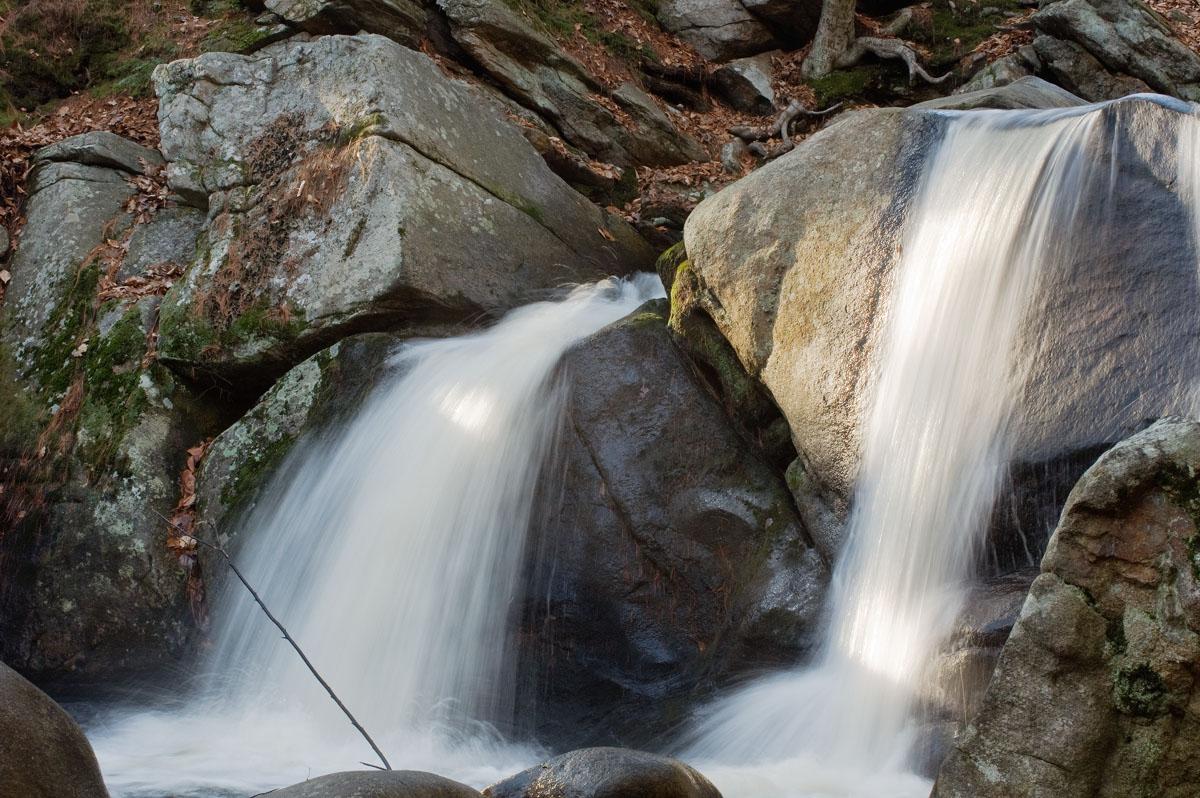 Sunlit waterfall flowing over large boulders in a natural, rocky landscape.
